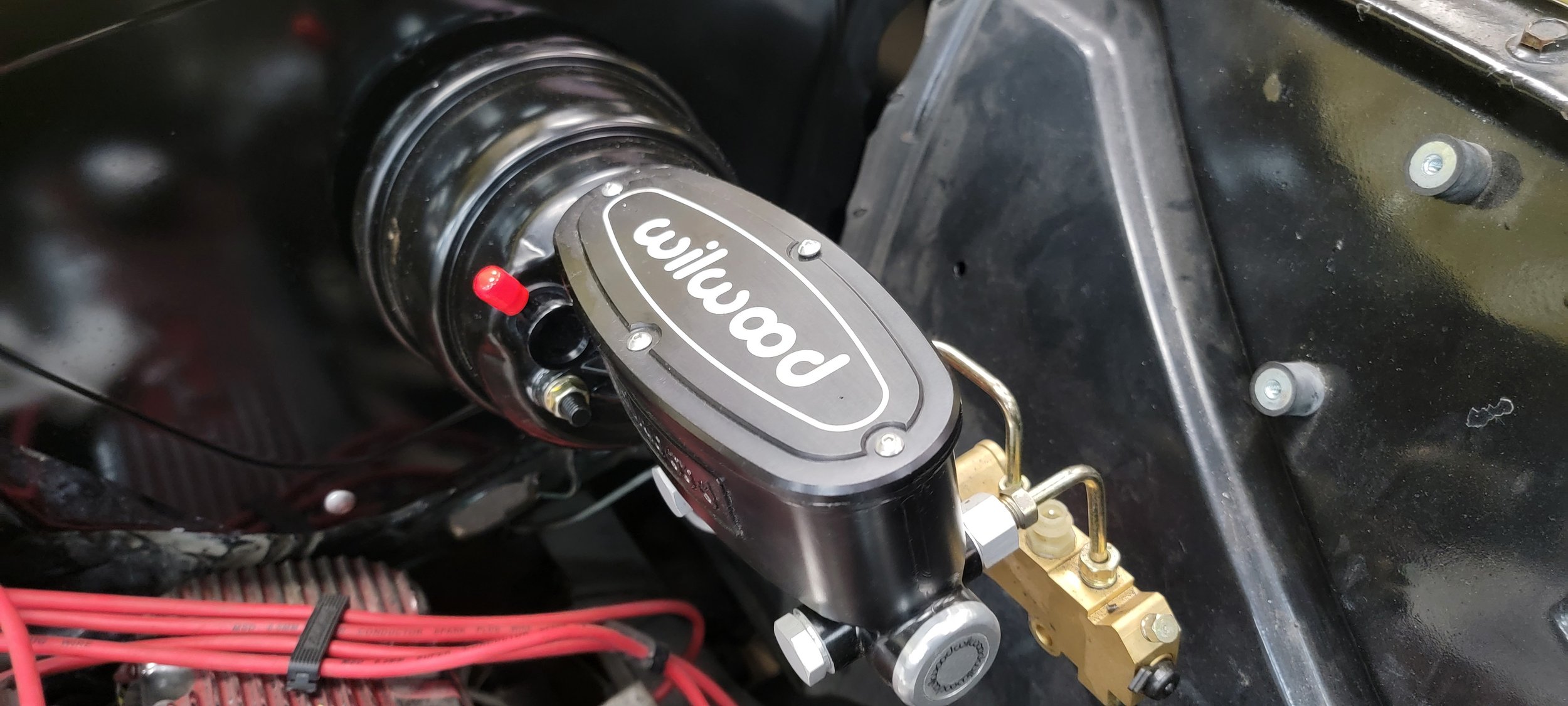 Close-up of a Wilwood brake master cylinder and brake system in a vehicle, with red suspension components and black engine parts visible.