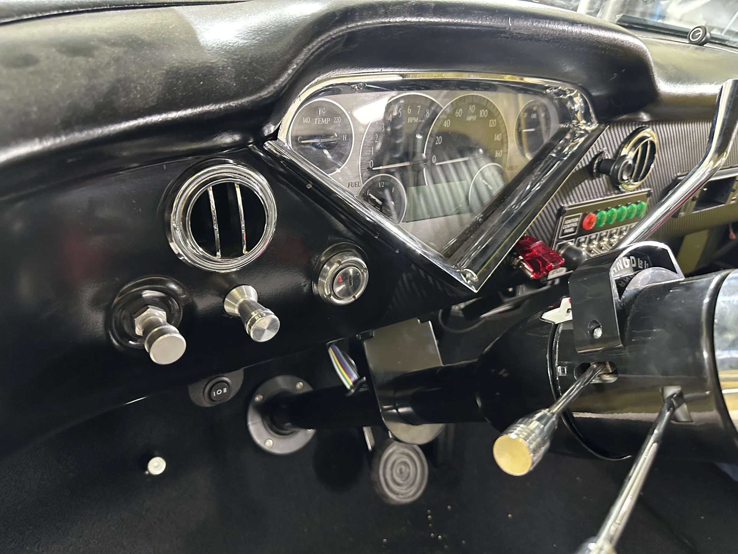 Close-up of a vintage car dashboard displaying gauges and controls, with speedometer, tachometer, fuel gauge, and switches, along with a vent and part of the steering wheel.