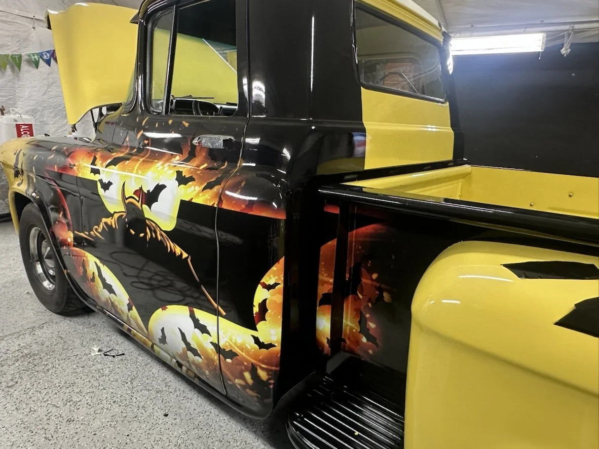 A vintage truck with a Halloween-themed paint job featuring a black silhouette of a witch riding a broomstick, bats, and a full moon on a black and yellow background.