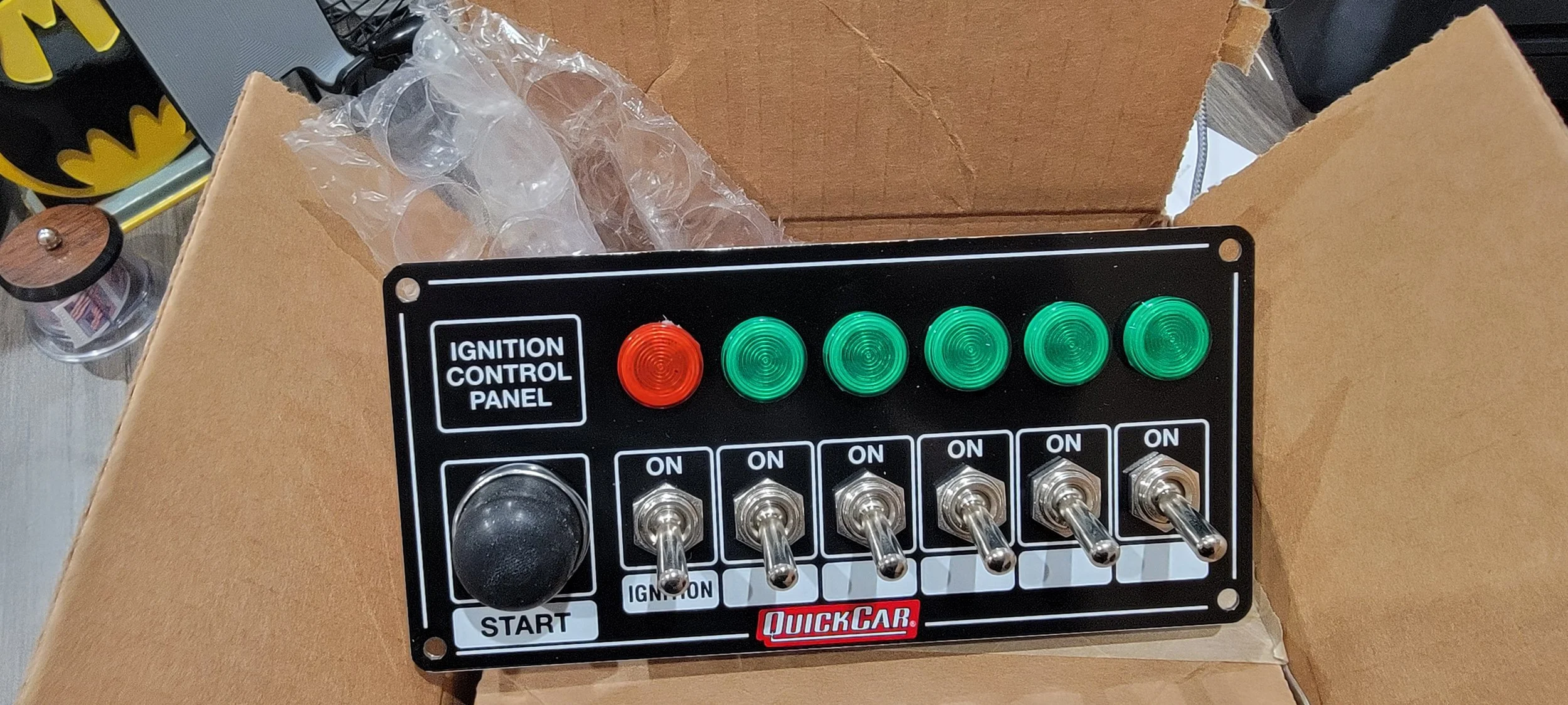 A black ignition control panel with red and green indicator lights, toggle switches, a start button, and a label reading 'IGNITION CONTROL PANEL,' placed on a cardboard surface.
