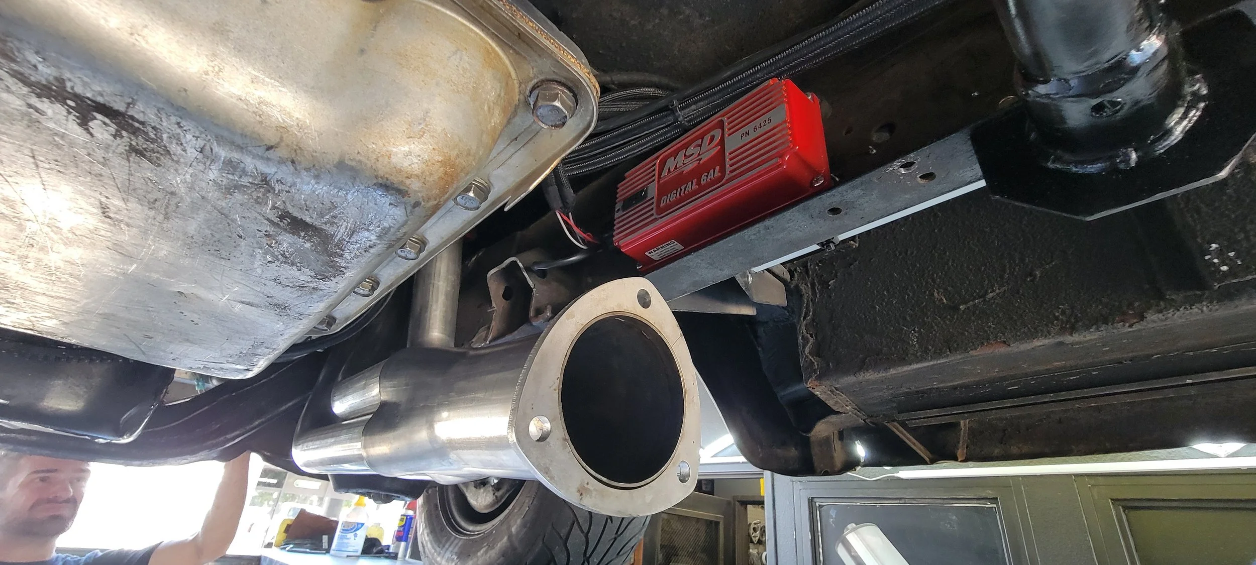 Underneath view of a car showing an aftermarket MSD digital ignition box installed on a black chassis, a metallic exhaust pipe, and a large metallic oil pan with bolts.