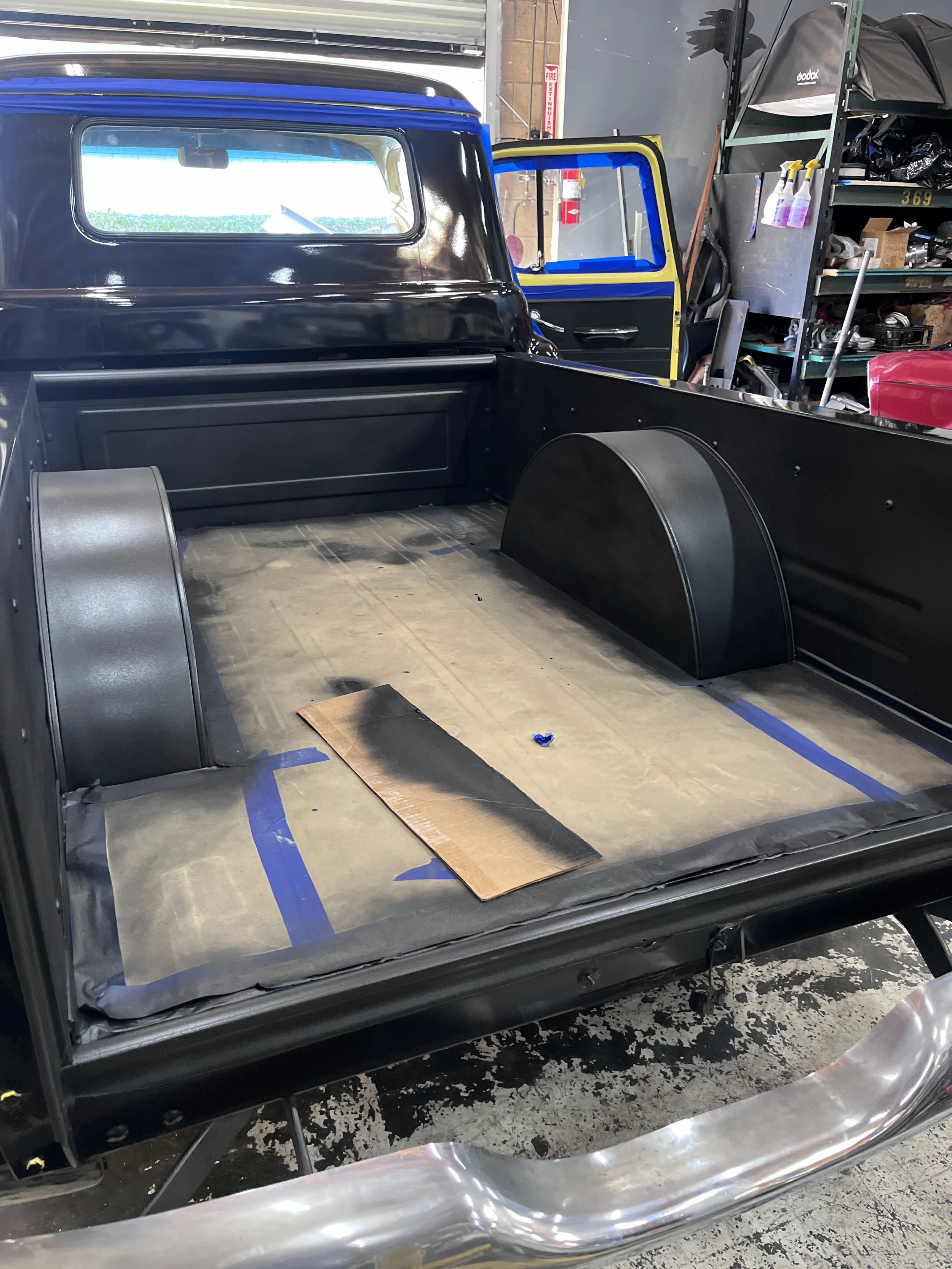 Black vehicle bed with fenders and wheel wells, possibly undergoing restoration or modification, inside a workshop with shelves and tools.