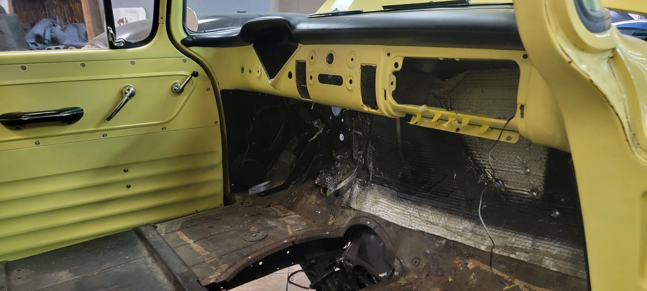 Interior of an old yellow vehicle in the process of restoration with exposed dashboard and floor, missing seats, and visible wiring.