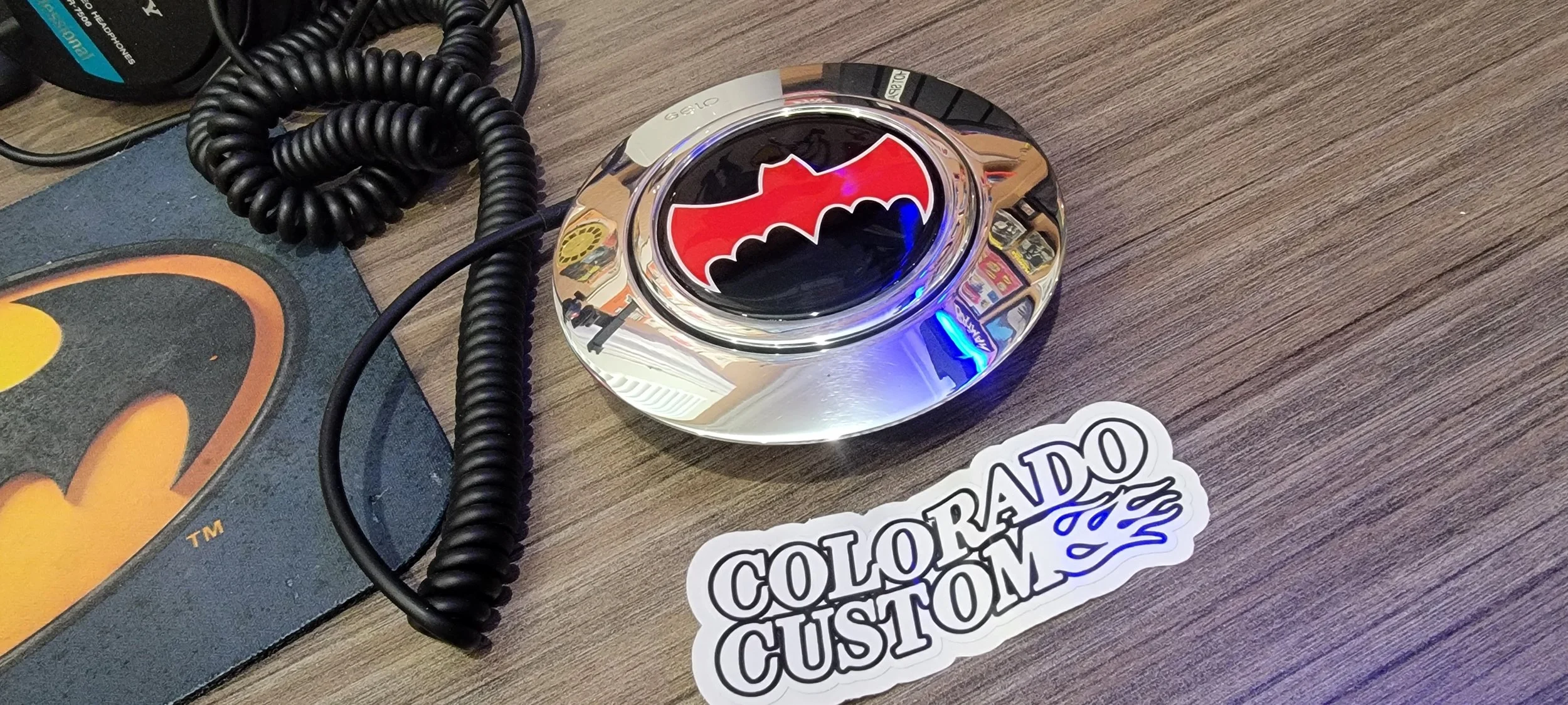 A round chrome-colored object with a Batman logo symbol on top, placed on a wooden surface. There is a black coiled telephone wire nearby and a sticker reading 'COLORADO CUSTOM' in the foreground.