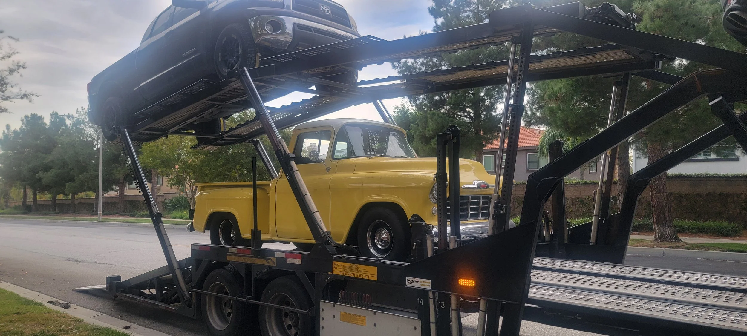 A flatbed tow truck carrying a yellow vintage pickup truck with a model of a black car on the top level.