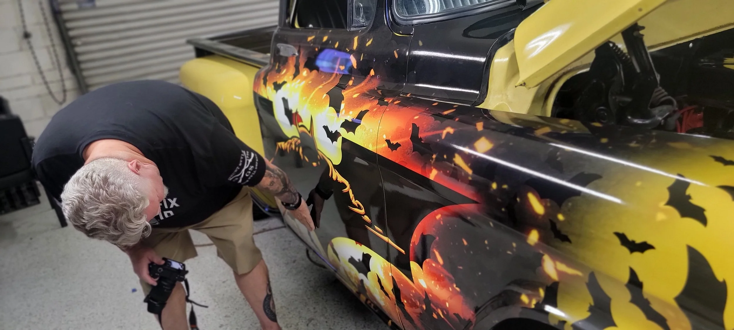 Person applying Halloween-themed fire and bats design to a black and yellow vehicle in a garage.