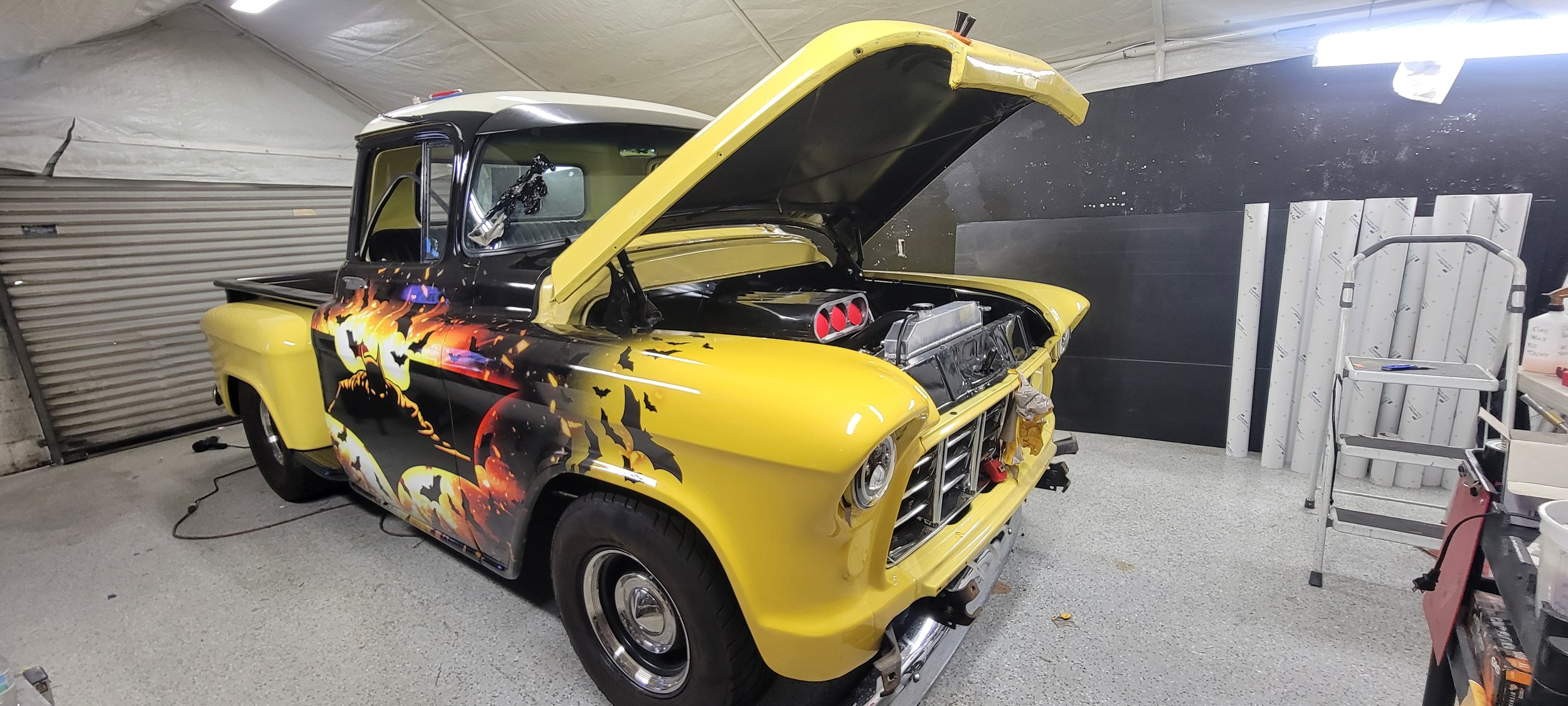 A yellow vintage pickup truck with its hood open, featuring Halloween-themed artwork of black bats, pumpkins, and a haunted house painted on the side, inside a garage with tools and equipment nearby.