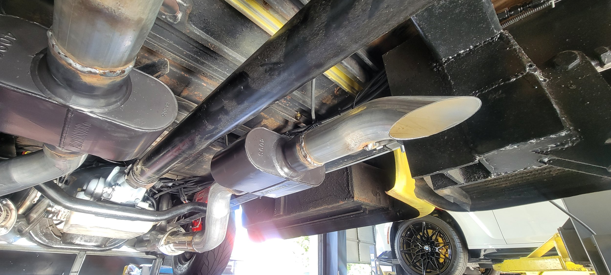 Underneath view of a car's aftermarket exhaust system, showing a polished stainless steel muffler and exhaust pipe, with black structural components and some yellow safety markings in the background.