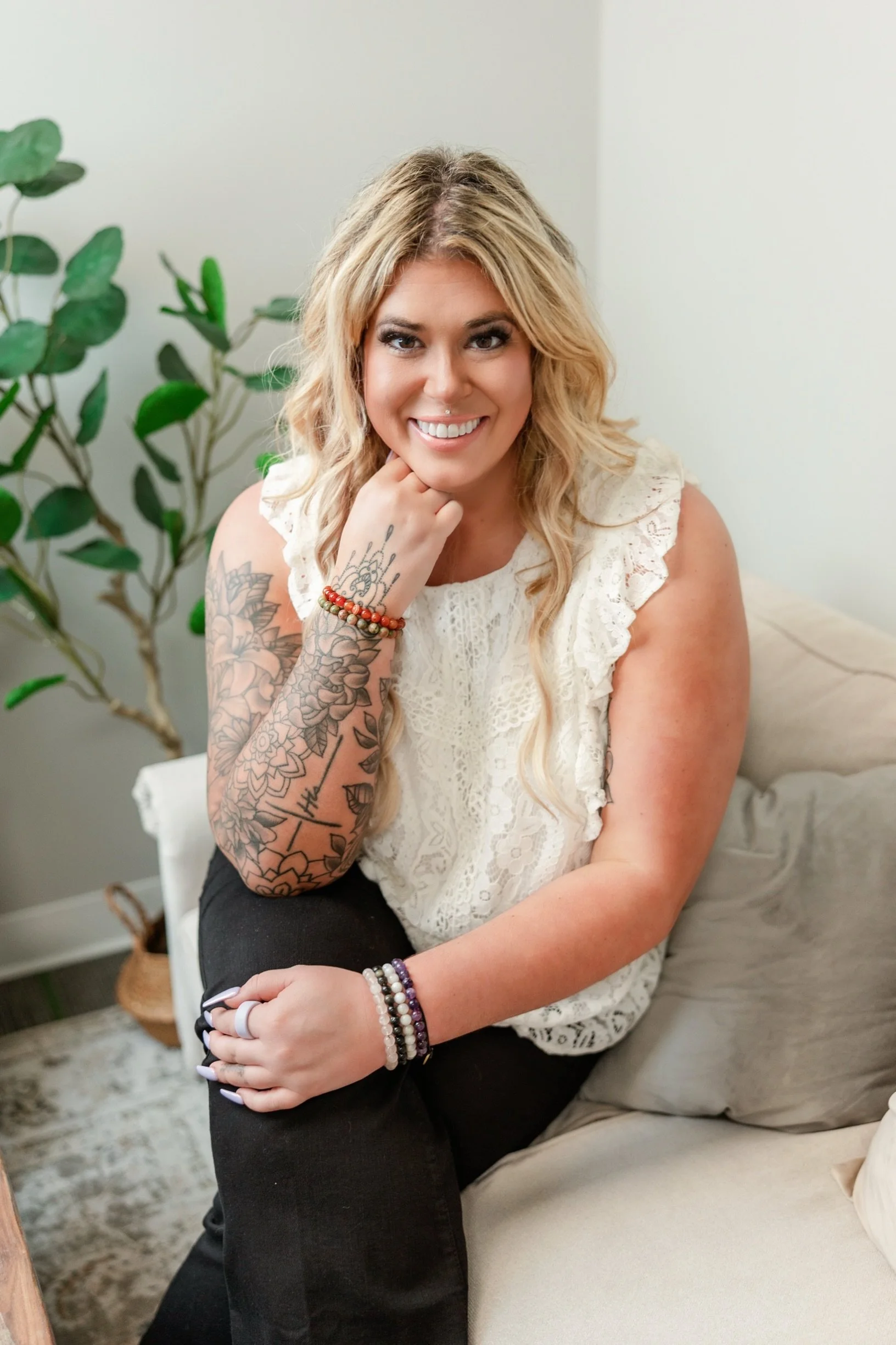 A woman with long blonde curly hair sitting on a beige couch, smiling at the camera, wearing a white lace top, multiple bracelets on her wrists, and tattoos on her left arm.
