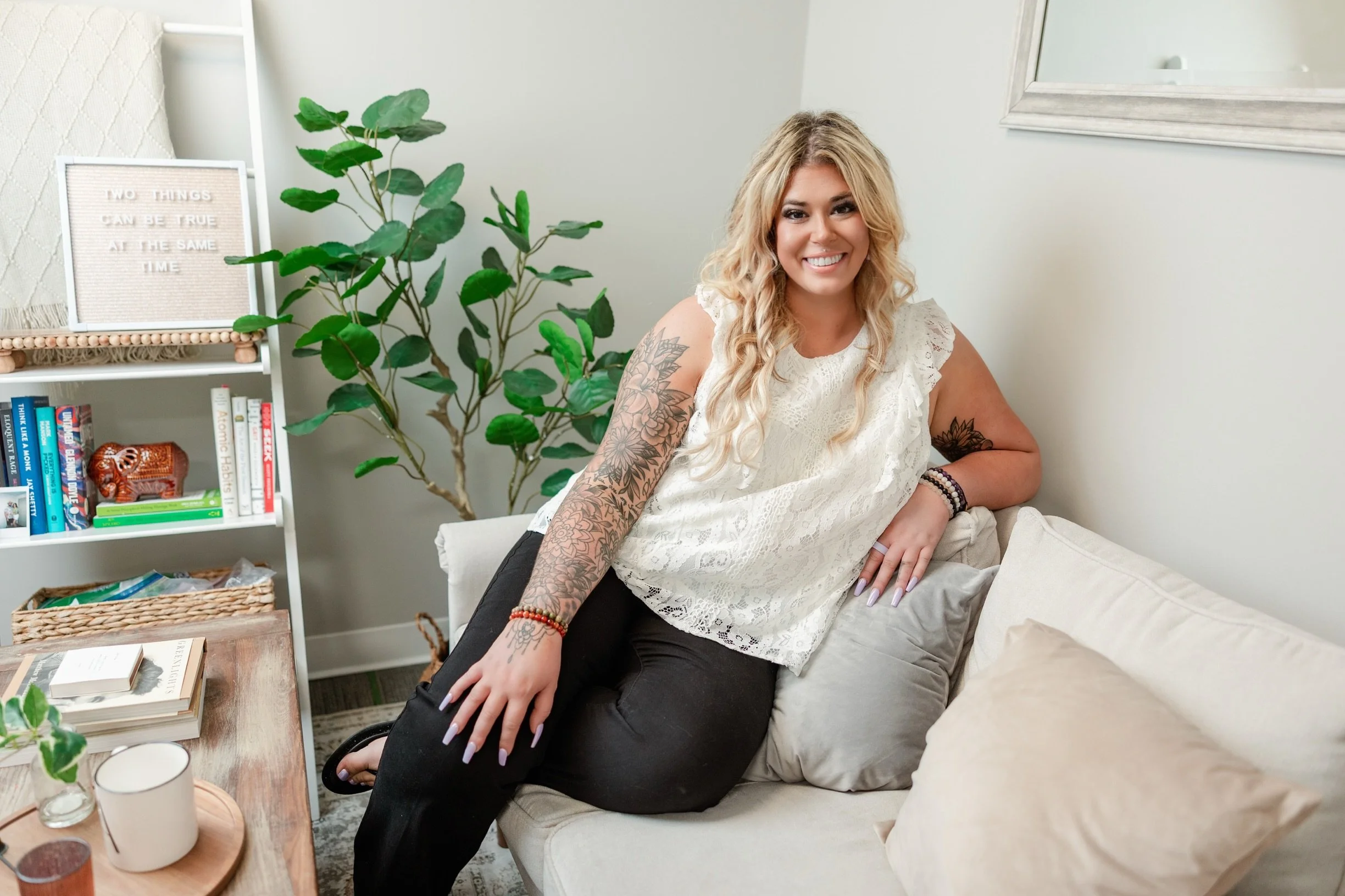A smiling woman with long curly blonde hair, tattoos on her arms, sitting on a beige couch in a room with a white wall, a green plant, and a bookshelf with books and a small picture frame.