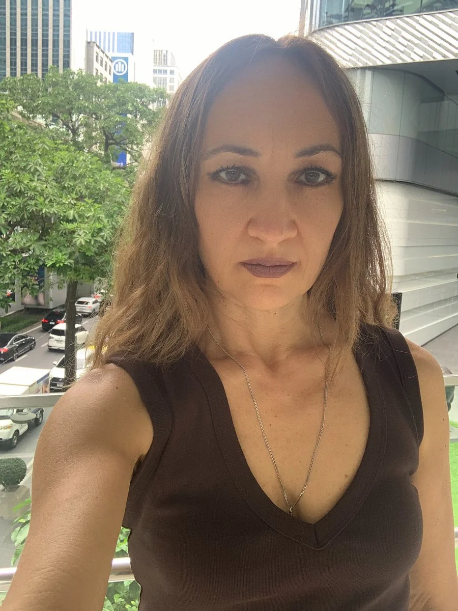 A woman at the cityscape background. She has wavy light brown hair, brown eyes, and is wearing a brown top and a thin silver necklace.