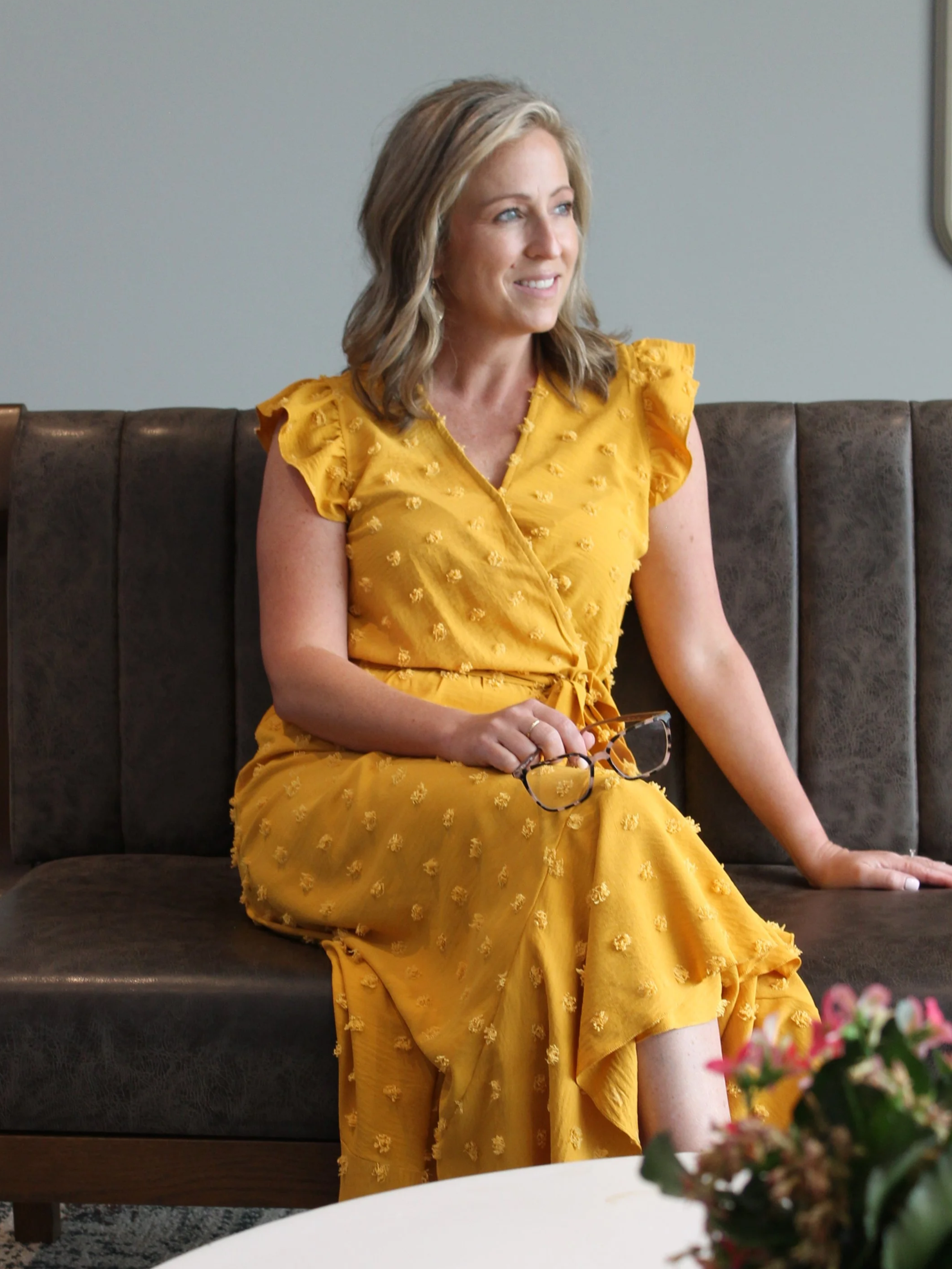A woman with blonde hair wearing a yellow dress with ruffled sleeves and embroidered flowers sits on a dark brown cushioned bench in a modern room, holding glasses and smiling while looking to her right.