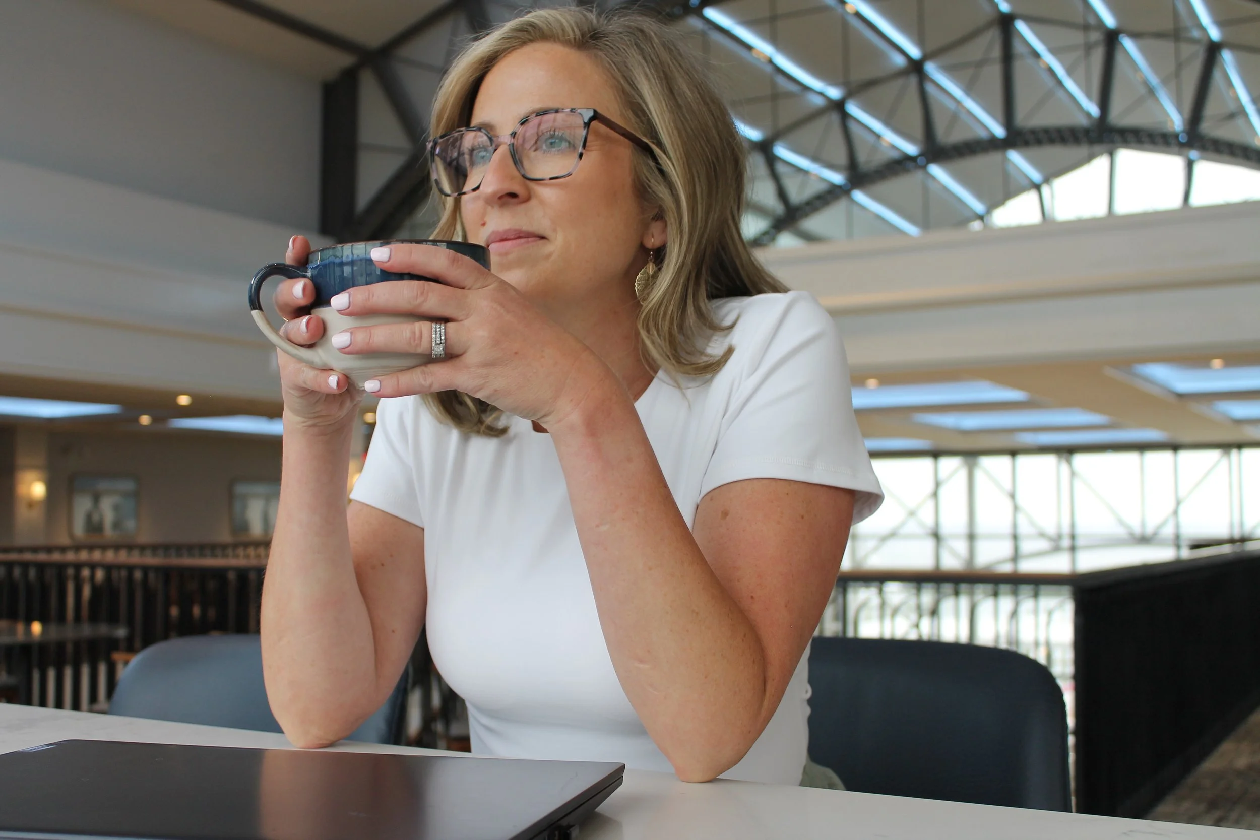 A woman with glasses and a white shirt sitting at a table in an indoor space, holding a mug close to her mouth, with a modern architectural ceiling in the background.