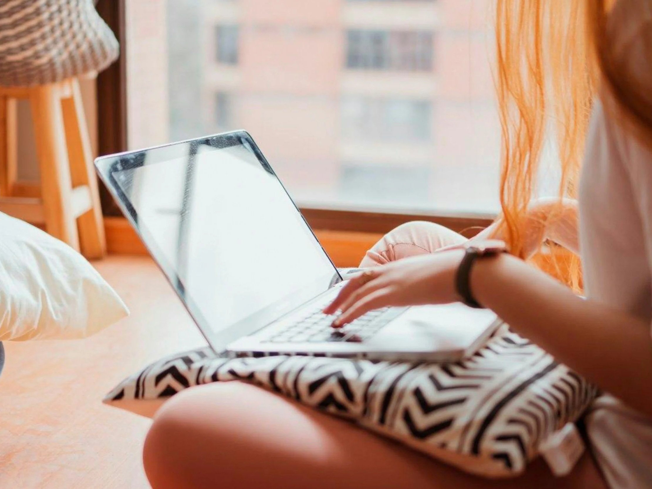 Person using a laptop computer near a window, wearing a patterned skirt or shorts, with a smartphone in hand, sitting on a bed with an orange curtain in the background.
