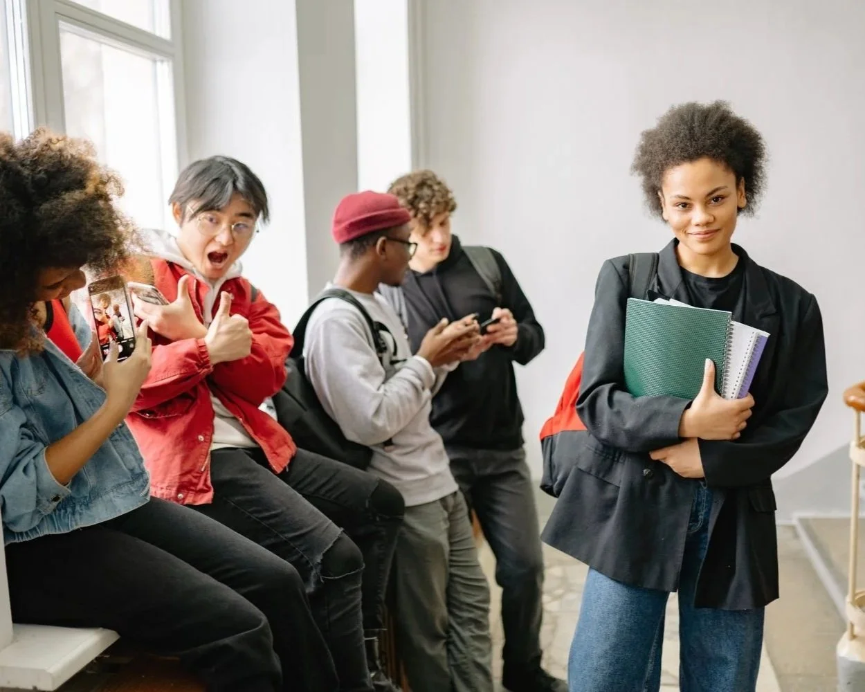 Group of five students, four sitting on a bench and one standing, inside a school hallway. The girl standing in front is holding notebooks and wearing a black jacket with a backpack, while the students on the bench are engaged with their phones.