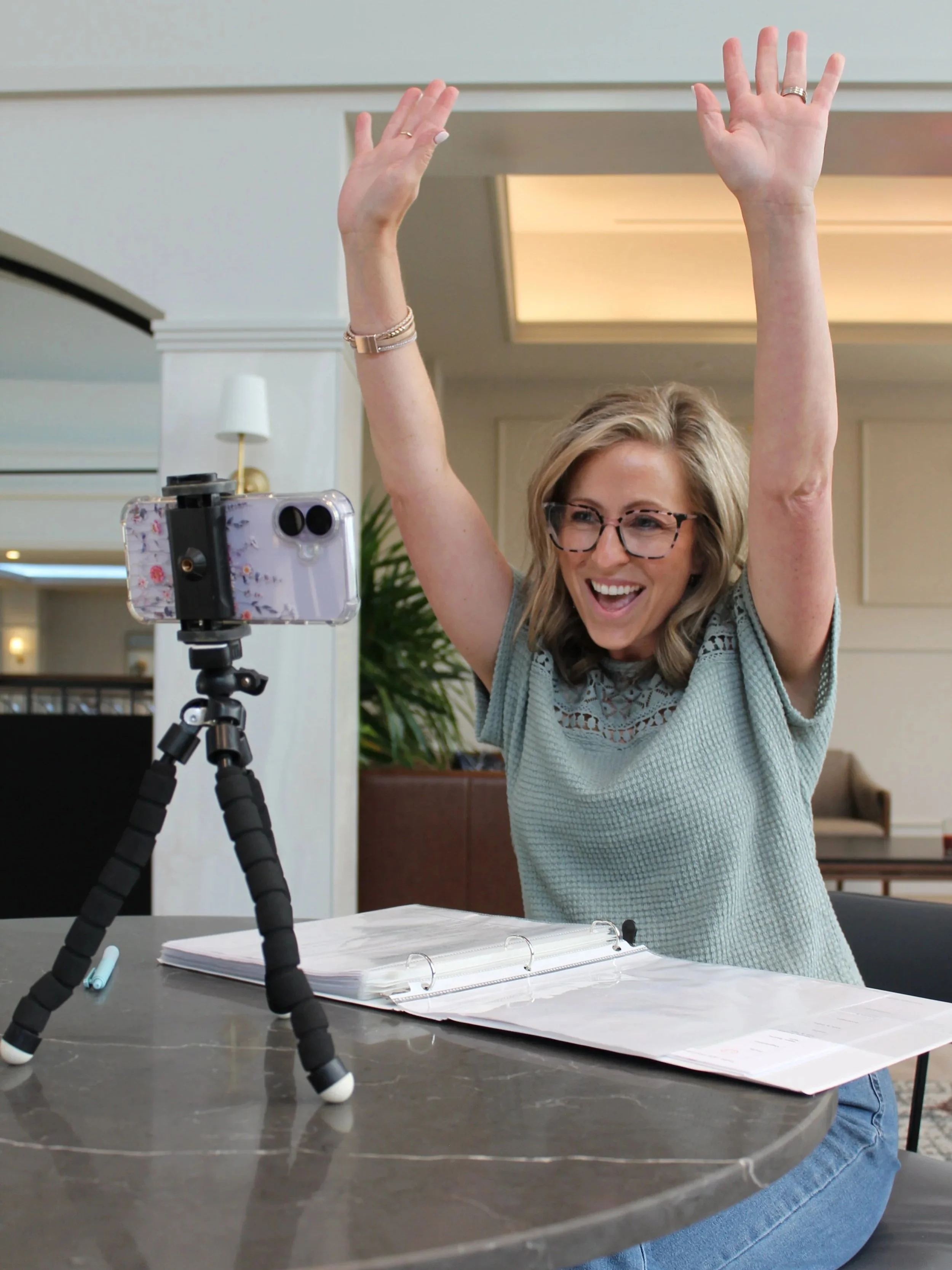 A woman with glasses and blonde hair standing at a table, raising her hands in celebration or excitement while filming herself with a smartphone on a tripod. The scene is set in a well-lit indoor space with papers and a notebook on the table.