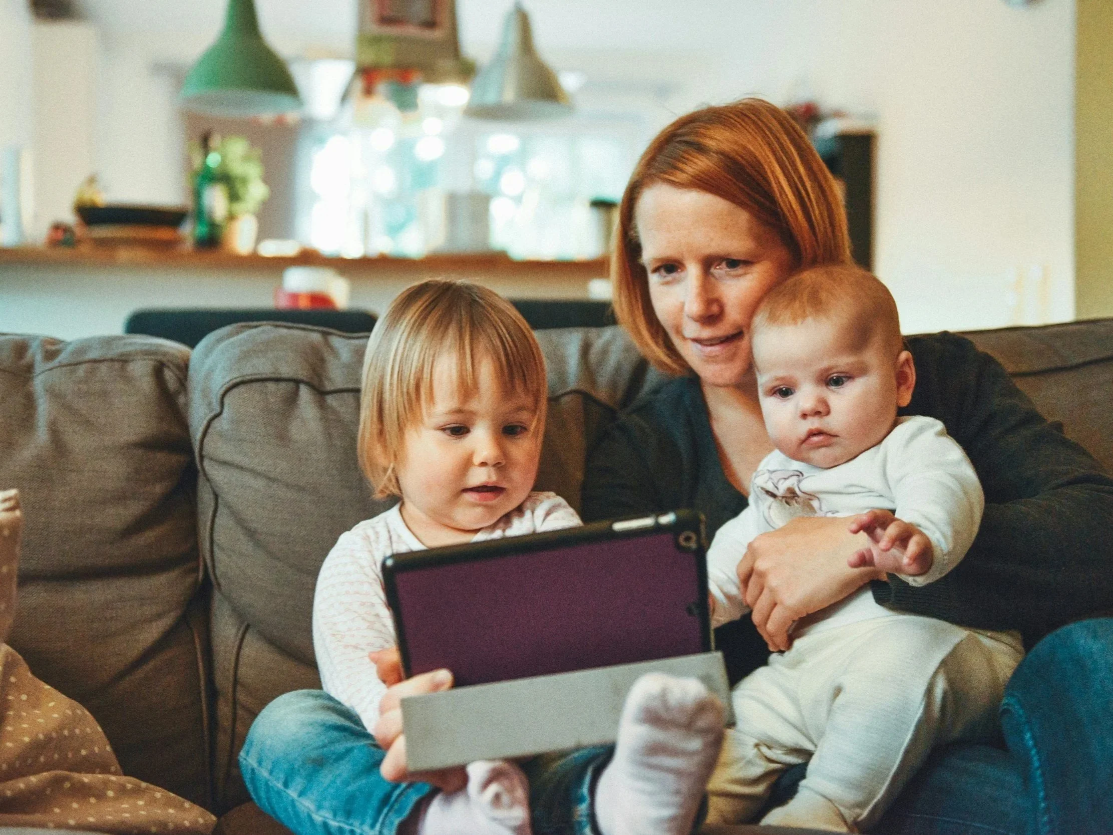 A woman with red hair and two young children sitting on a couch, looking at a tablet device together. The children have light brown hair and are wearing light-colored clothing. The background shows a home interior with hanging lights and kitchen items.