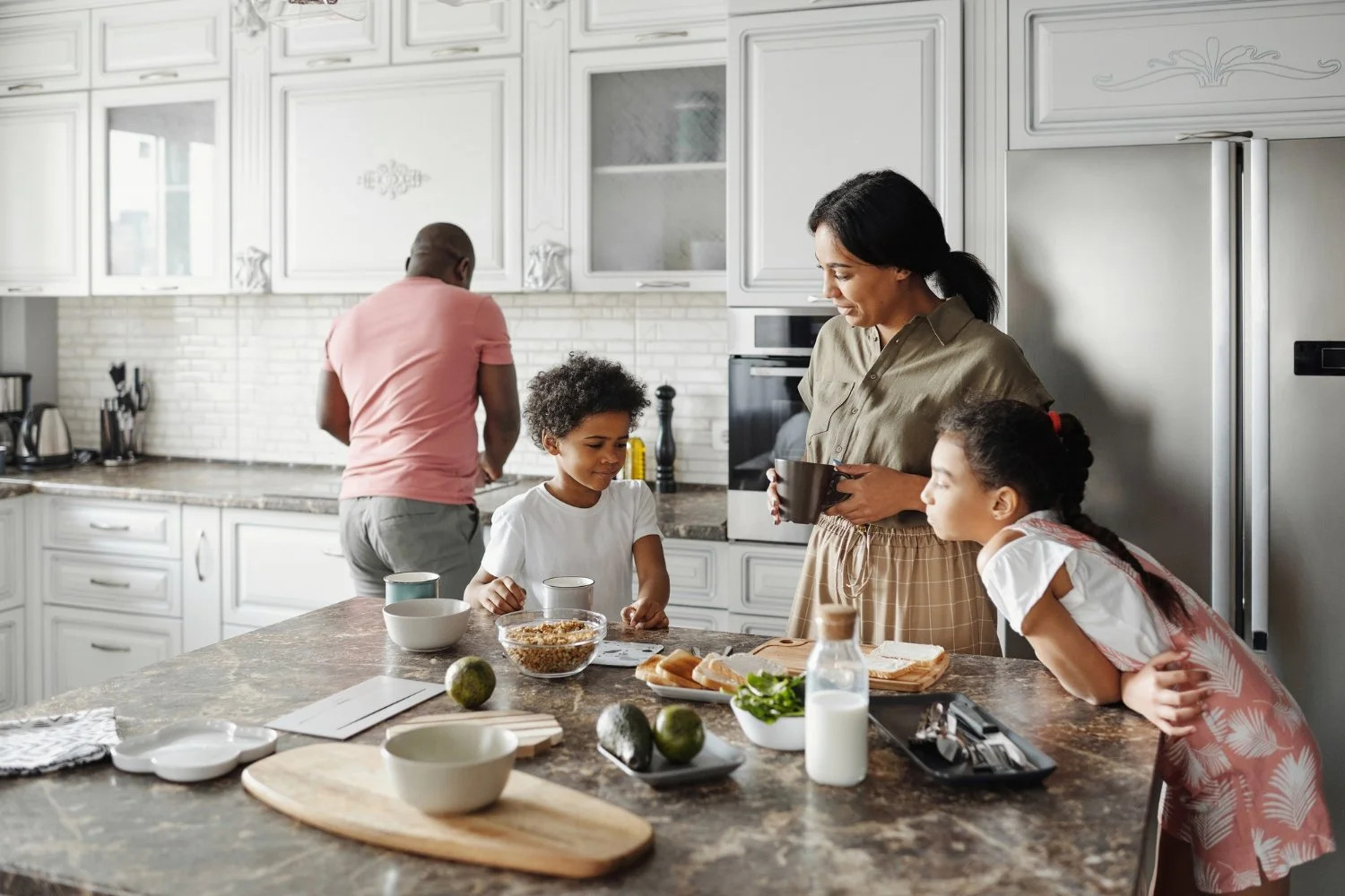 A family in a bright kitchen preparing breakfast together. There are two children, a woman, and a man. The woman is holding a bowl and the children look interested. The kitchen has white cabinets and a marble countertop with bowls, bread, and avocados on it.