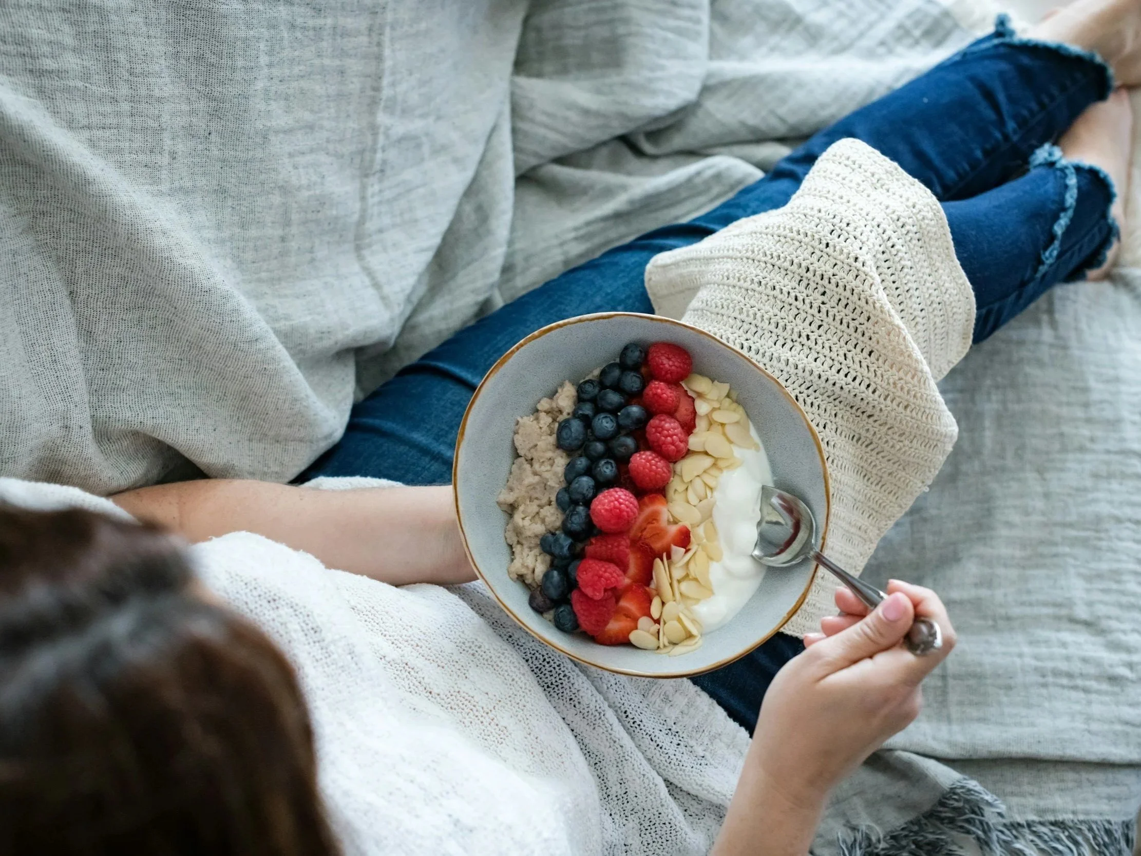Person sitting on a sofa with a gray shirt and ripped denim shorts, holding a bowl of yogurt topped with blueberries, raspberries, sliced strawberries, sliced almonds, and granola.