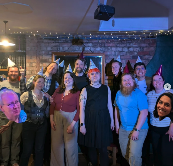 A group of people celebrating at a party with colorful cone party hats, in a cozy indoor setting with string lights and a brick wall. They are smiling and posing for the photo.