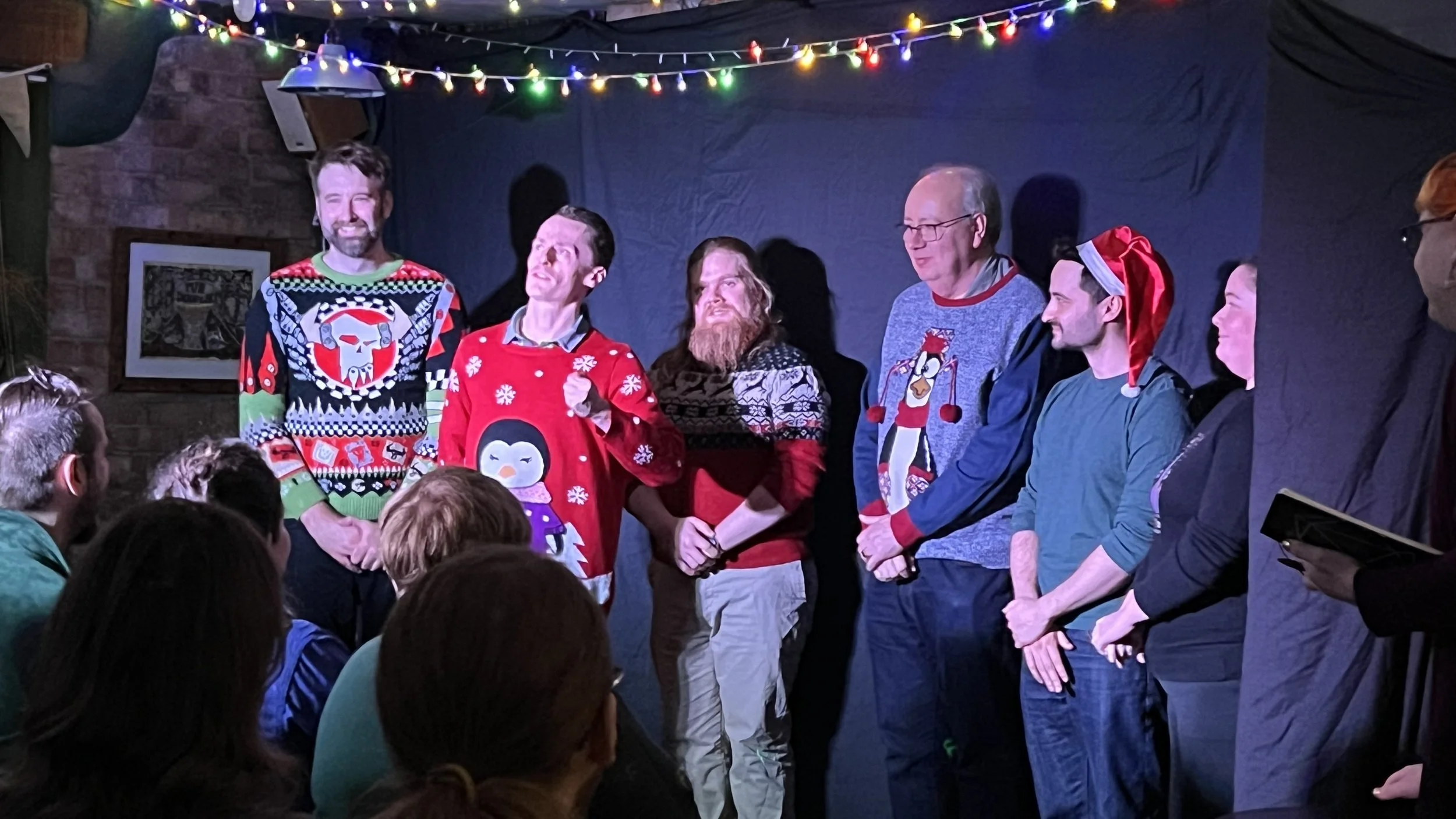 A group of people standing on a stage decorated with Christmas lights, celebrating in festive sweater and accessories, surrounded by seated audience members.