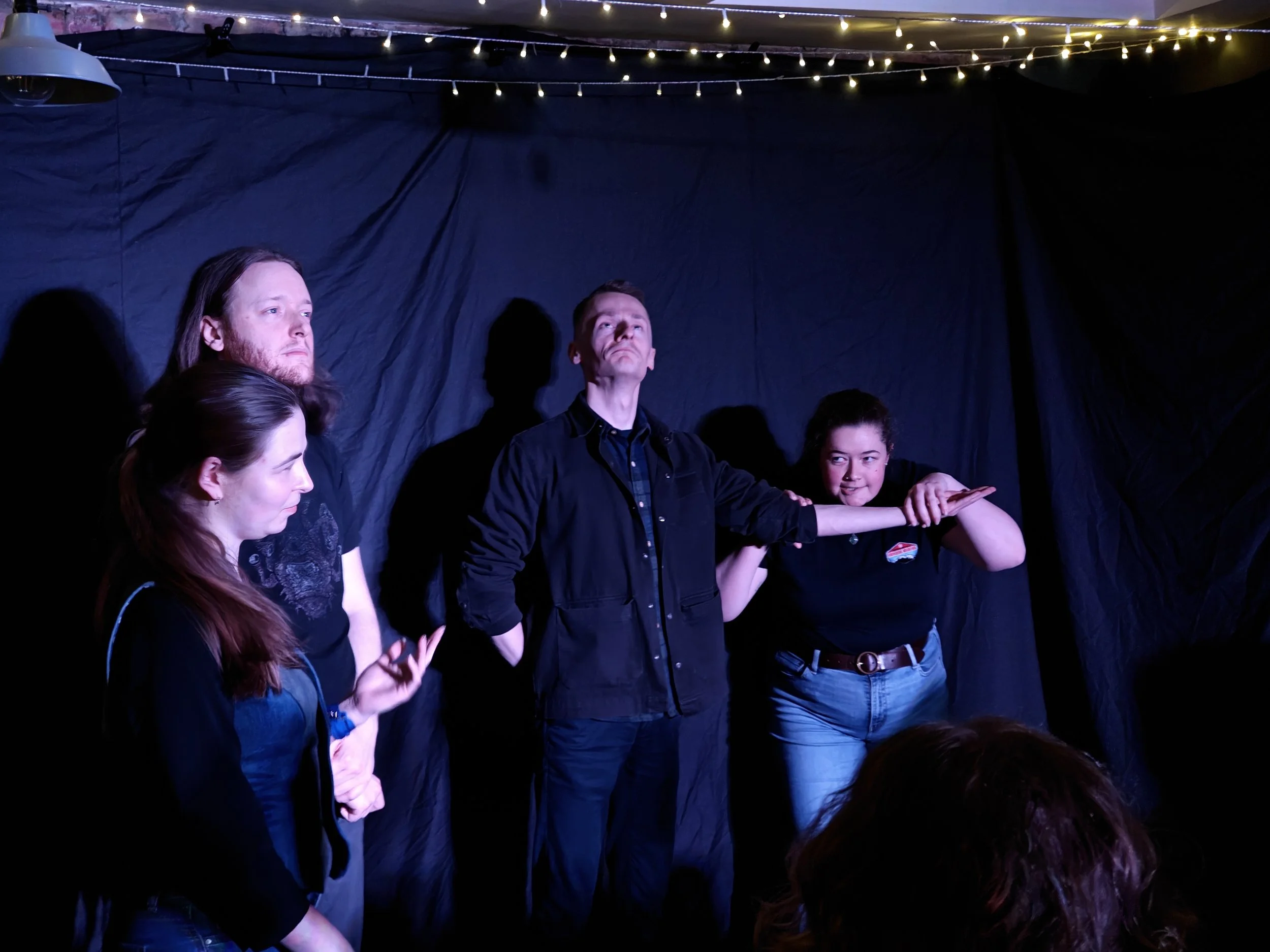 People standing in a semi-circle in a dimly lit room with string lights overhead, engaging in a conversation or activity.