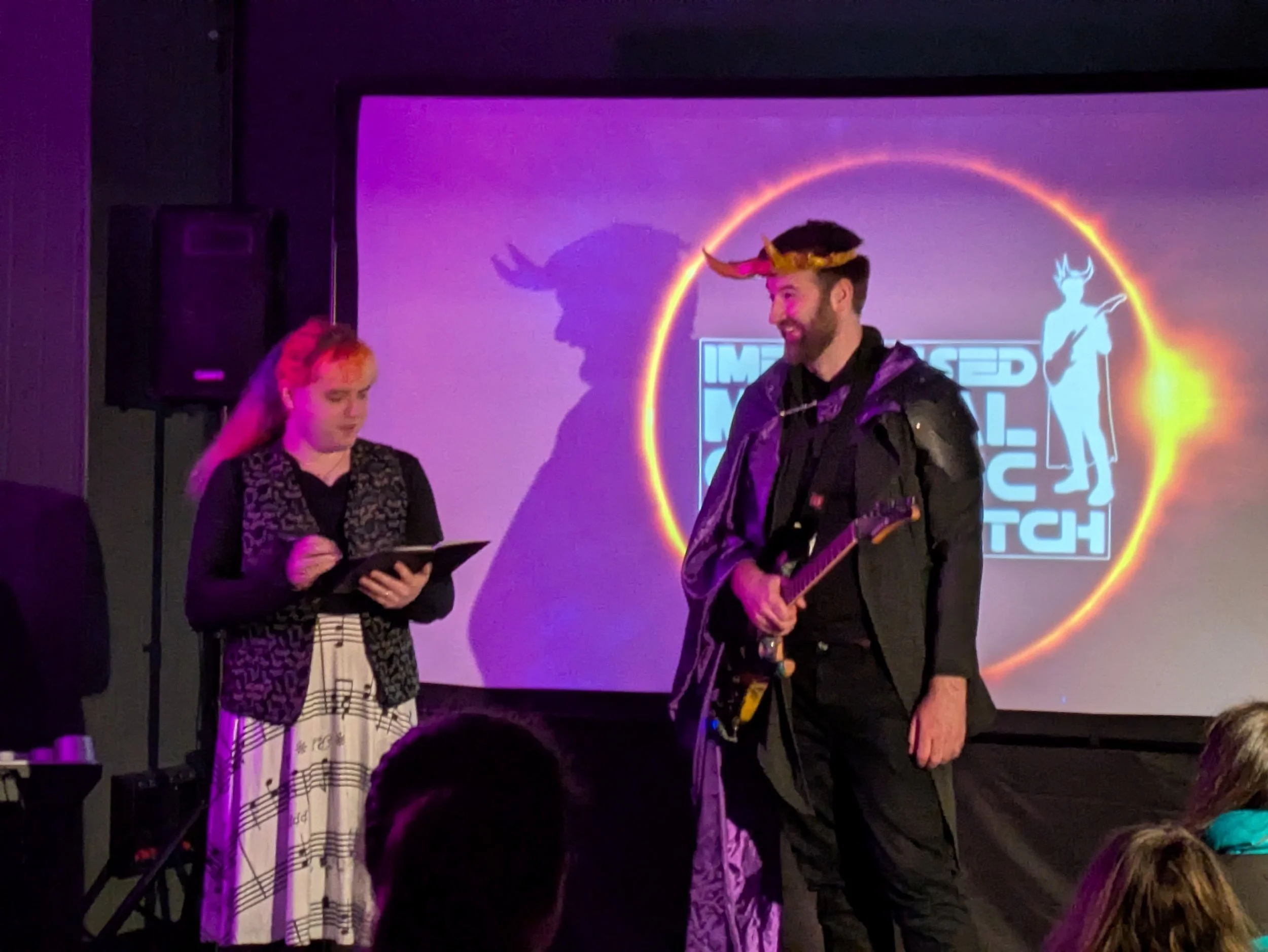 People performing on stage with a neon-lit backdrop, including a person wearing a headpiece with horns and holding a guitar.
