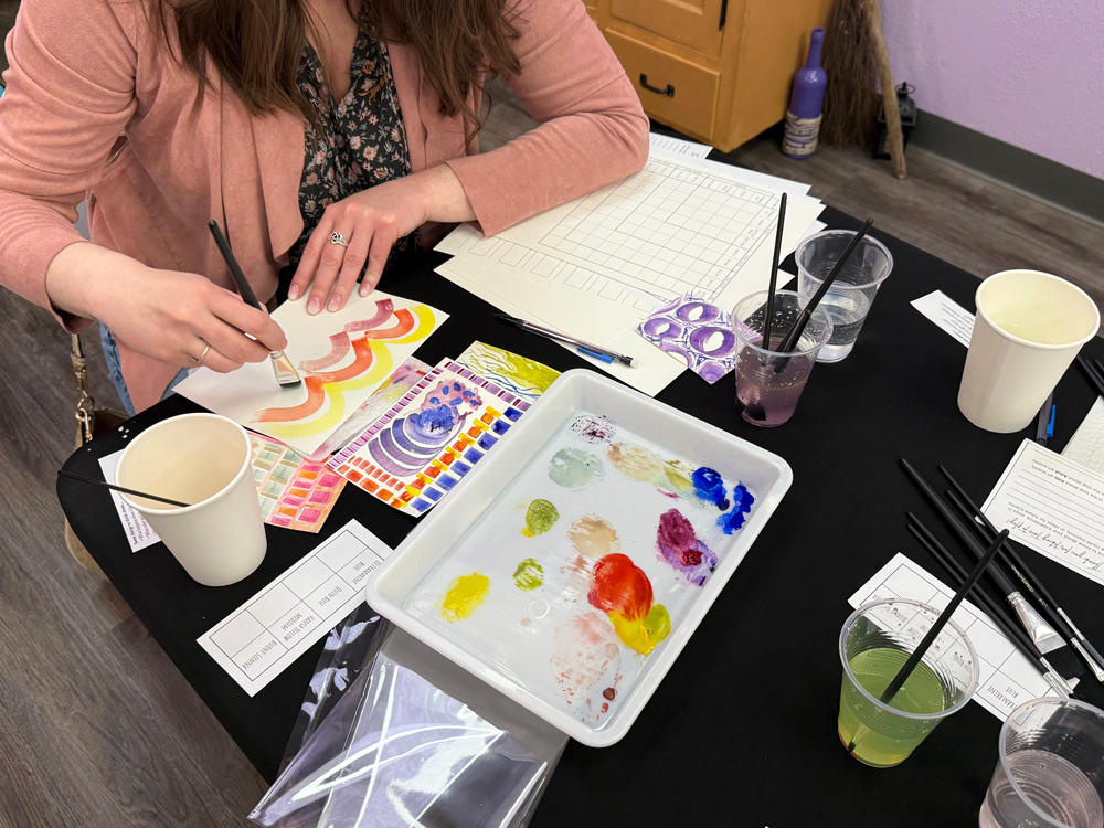 A woman painting abstract watercolor designs on paper at a table filled with painting supplies, cups of water, and color palettes.