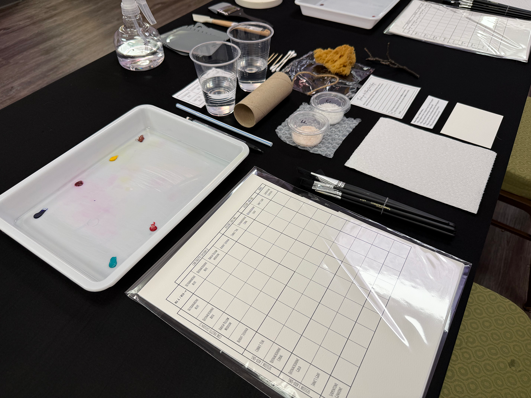 Table with paint palette, paint samples, water glasses, paintbrushes, paper, and art supplies.