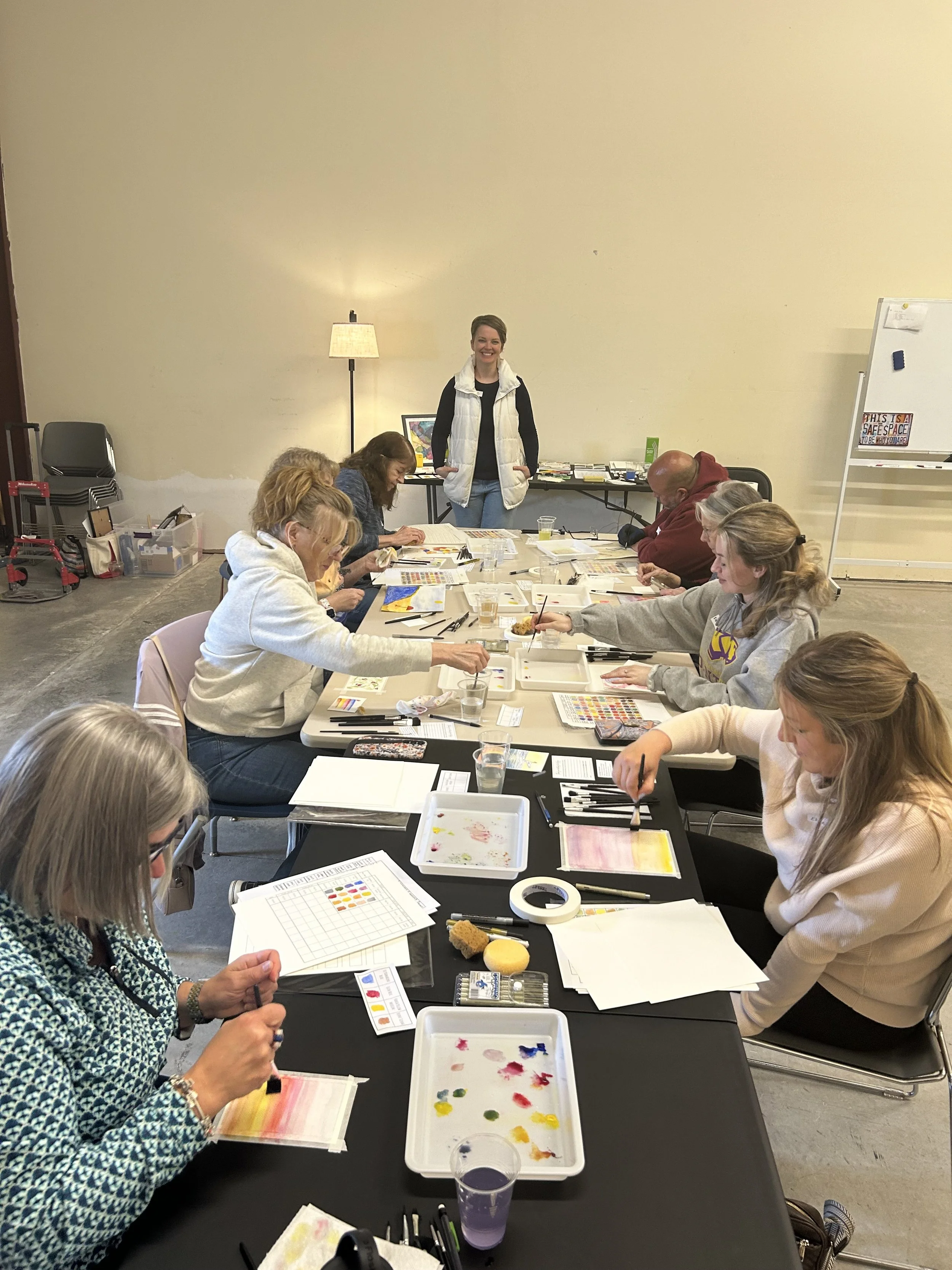 A group of people sitting around a long table painting watercolor artworks in a room with plain beige walls. There is a woman standing at the end of the table smiling at the camera.