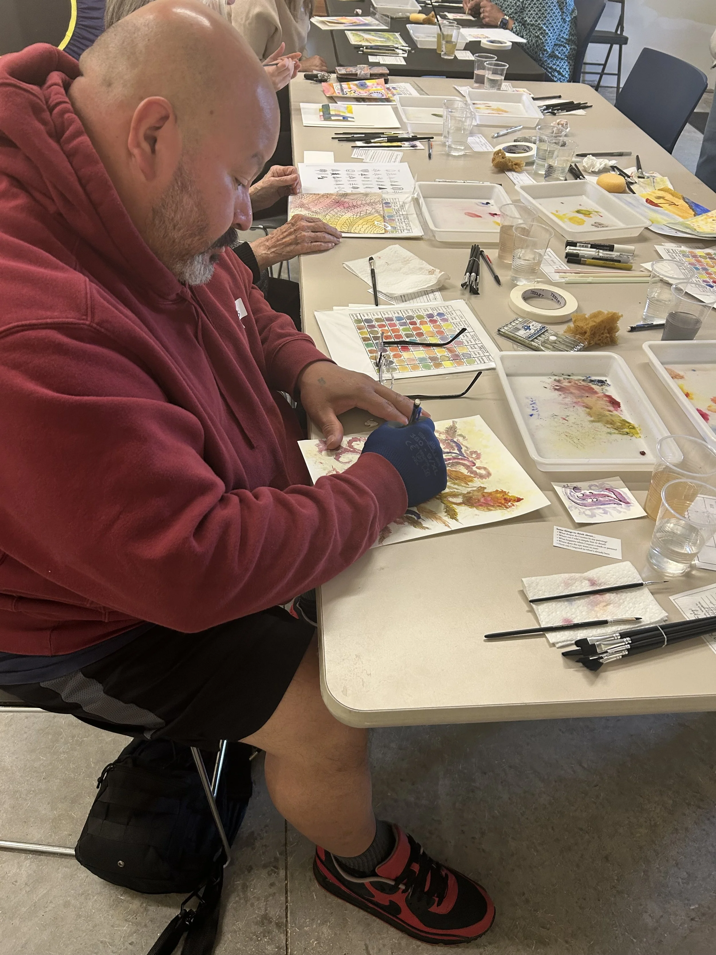 A person with a shaved head wearing a red hoodie and black shorts is sitting at a table and painting with watercolors. The table is filled with various watercolor supplies, brushes, trays with paint, pens, papers, and glasses of water. Other people a
