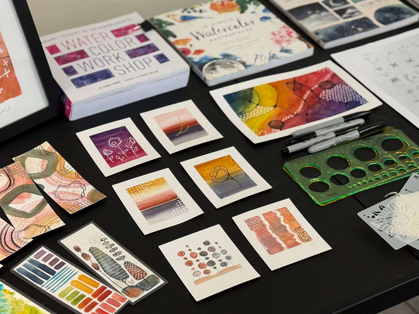 Table with watercolor art supplies and small watercolor paintings. There are various art books, colored markers, and a green stencil ruler on the table.