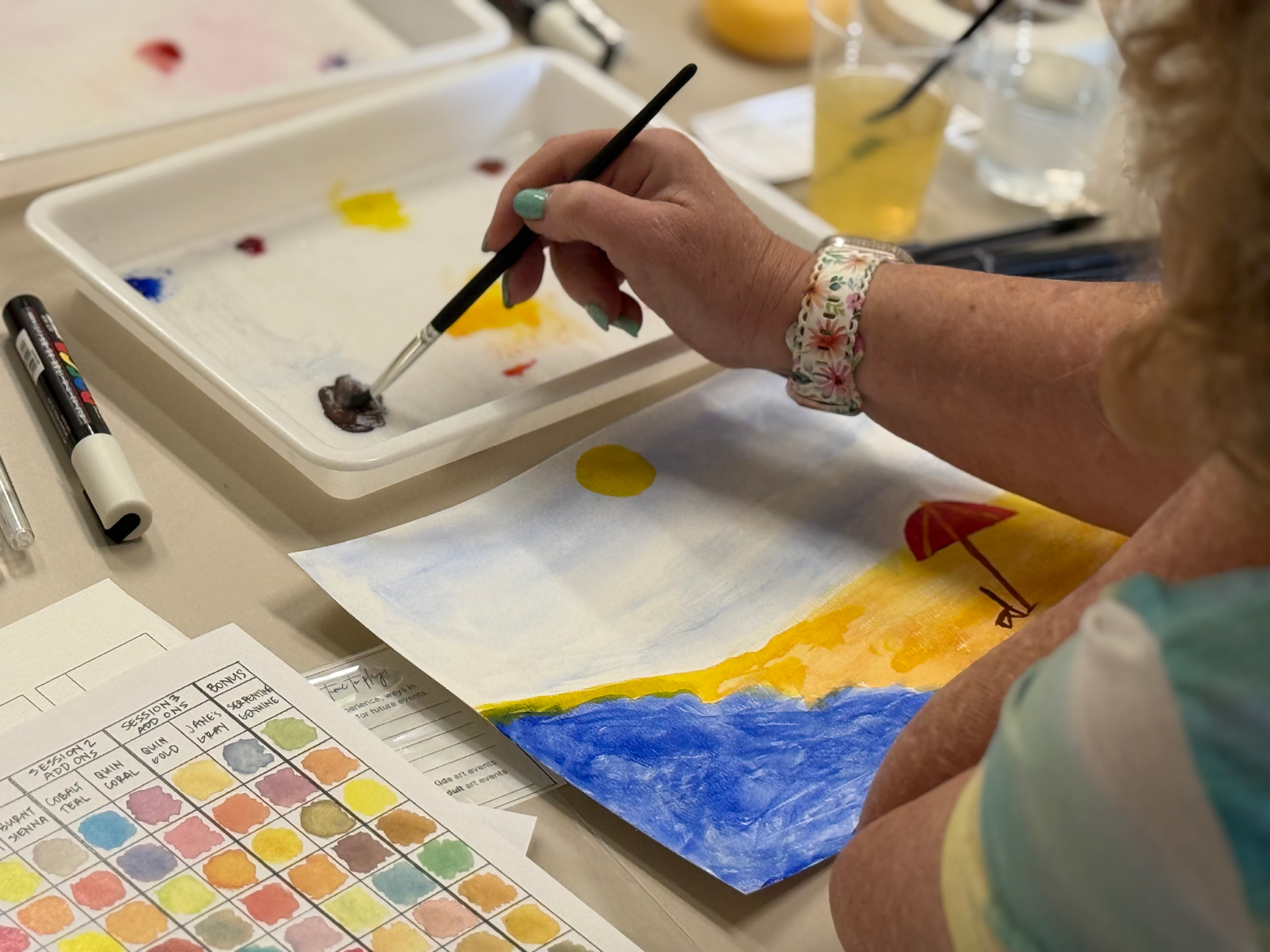Person painting with watercolors on paper, surrounded by color swatches and art supplies.