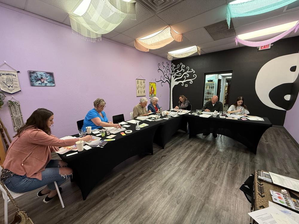 Group of six women and one man sitting at tables in a room with pastel and black walls, decorated with hanging fabric and a black-and-white mural of a yin-yang symbol and a tree.