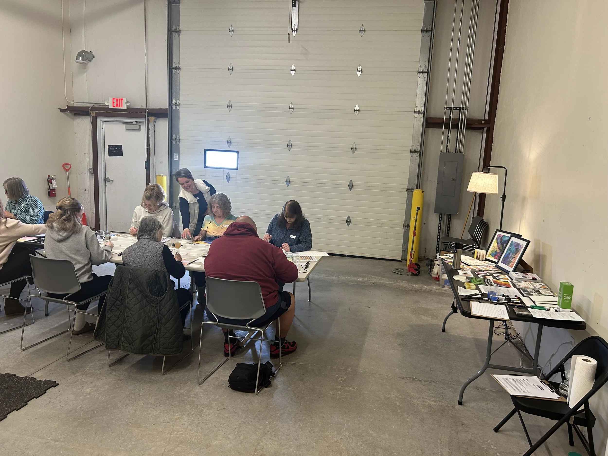 A group of people sitting around a table in an industrial-style room with a high ceiling, large garage door, and concrete floor. Some artworks and supplies are laid out on a table to the right, and a woman is standing smiling near the center, engagin