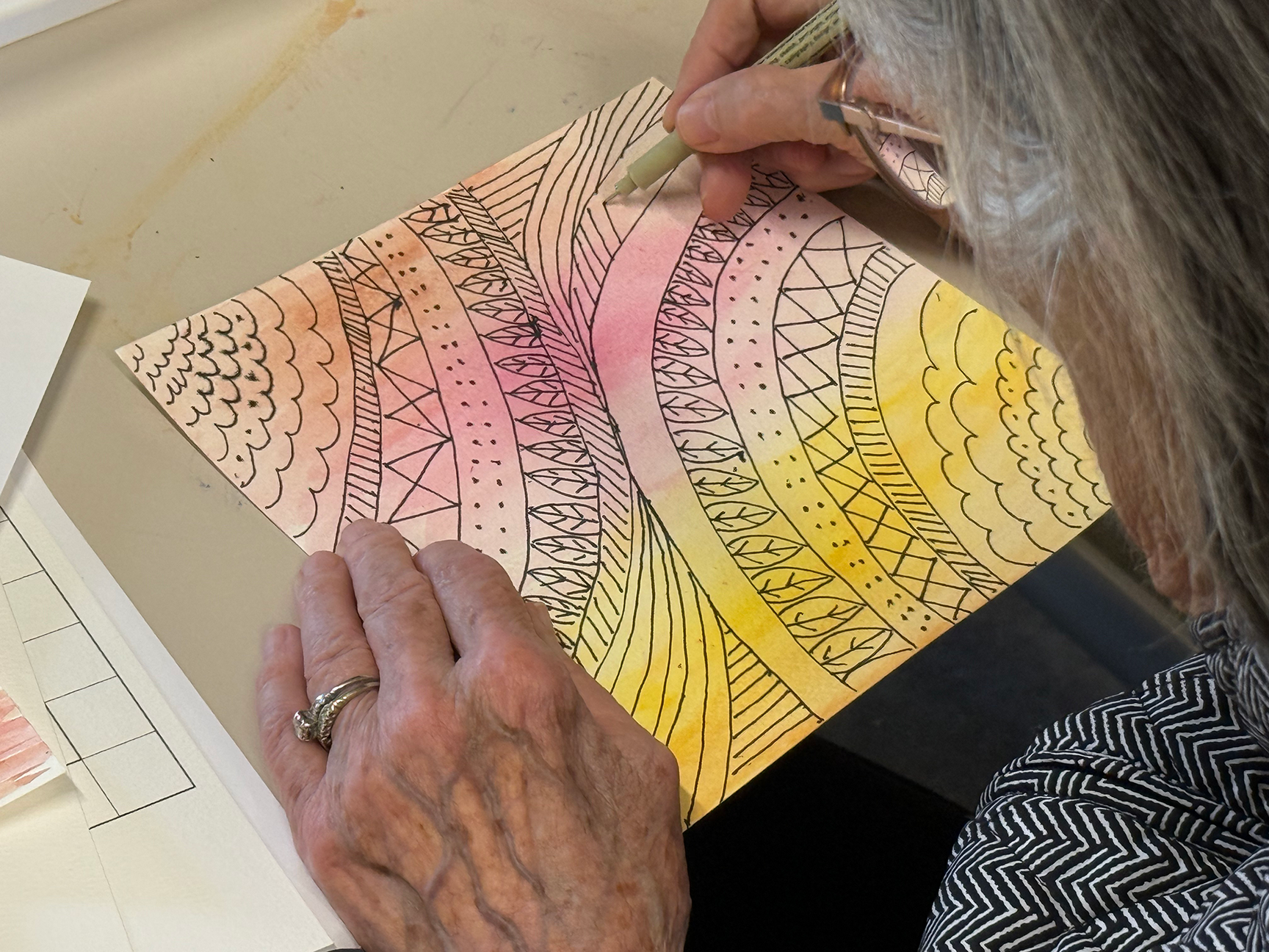 Older person drawing or coloring on a piece of paper with black patterns and a gradient of pink and yellow watercolor backgrounds.