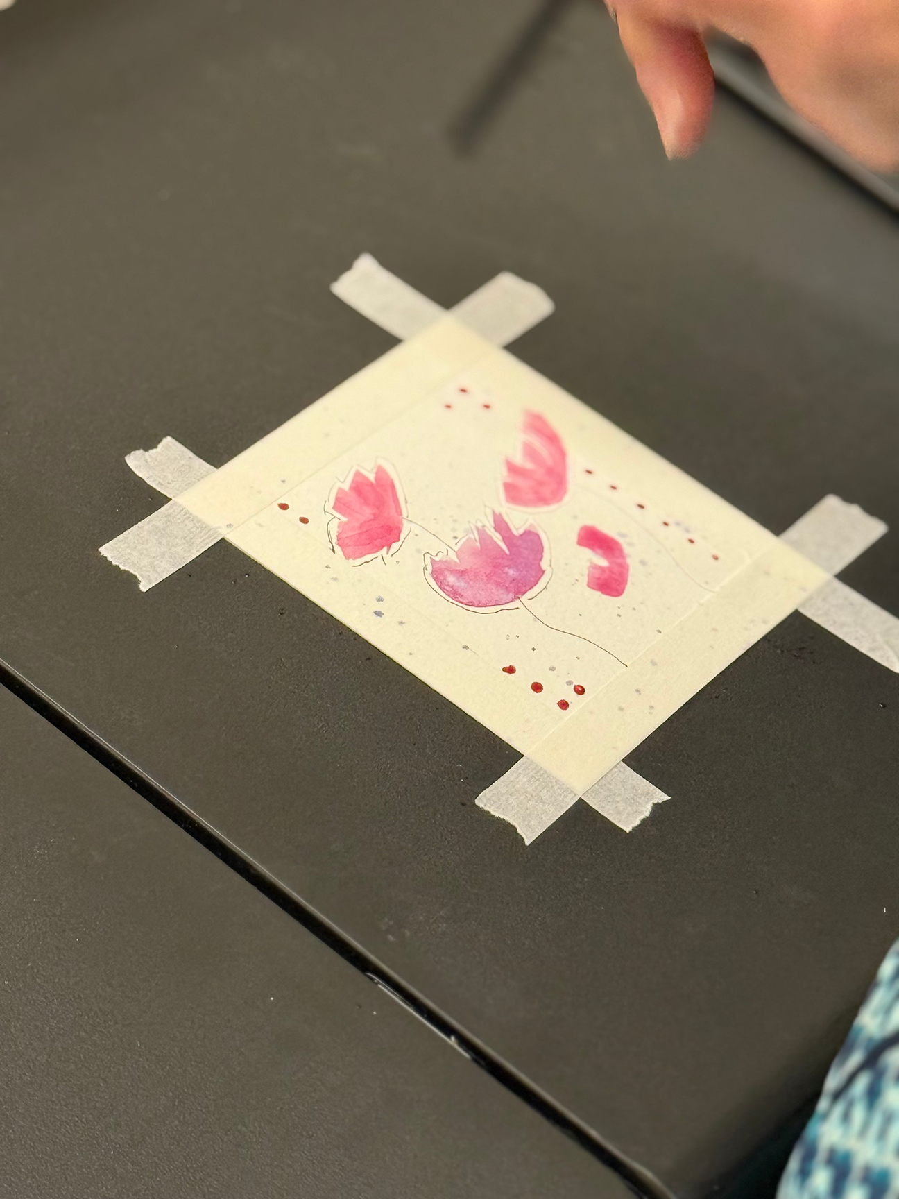 A piece of paper taped to a dark surface with masking tape, featuring a pink watercolor painting of three butterfly wings.