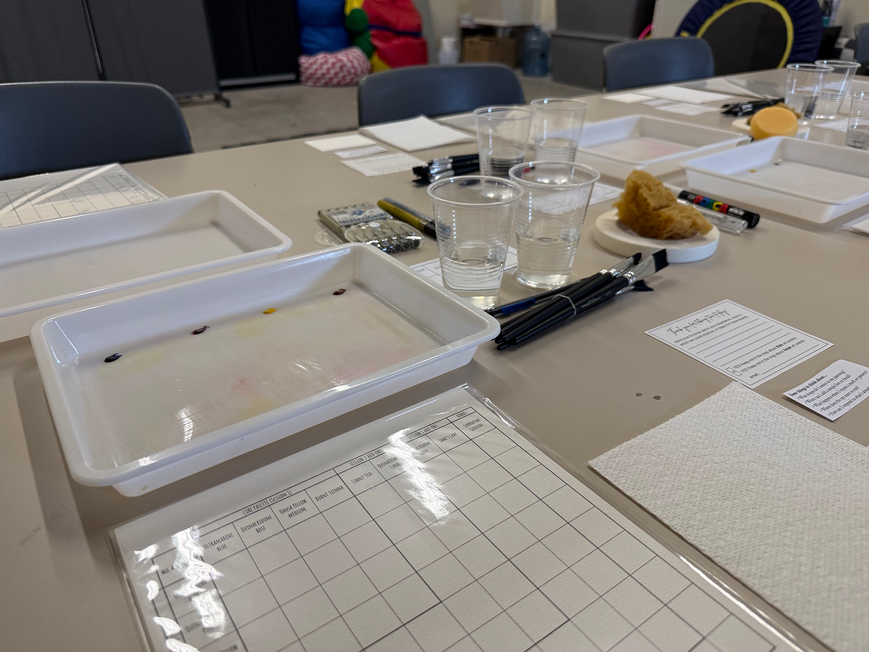 A table set up for a wine tasting, with empty tasting sheets, black pens, clear plastic cups filled with water, and small bread pieces on white plates.