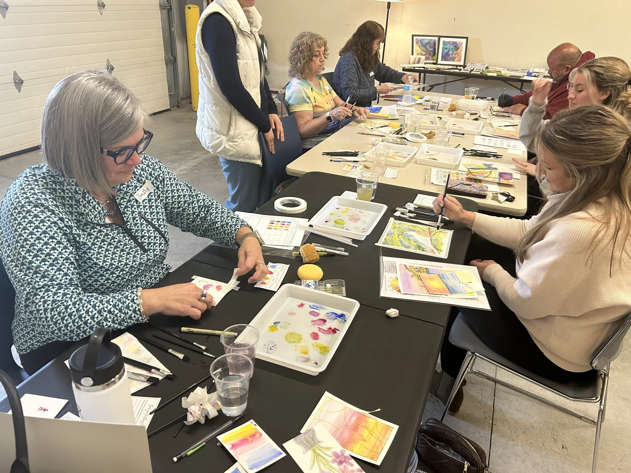 Group of women participating in an art class, painting watercolor pictures of flowers and nature at a long table with supplies like watercolors, brushes, and paper, in a room with a garage door.