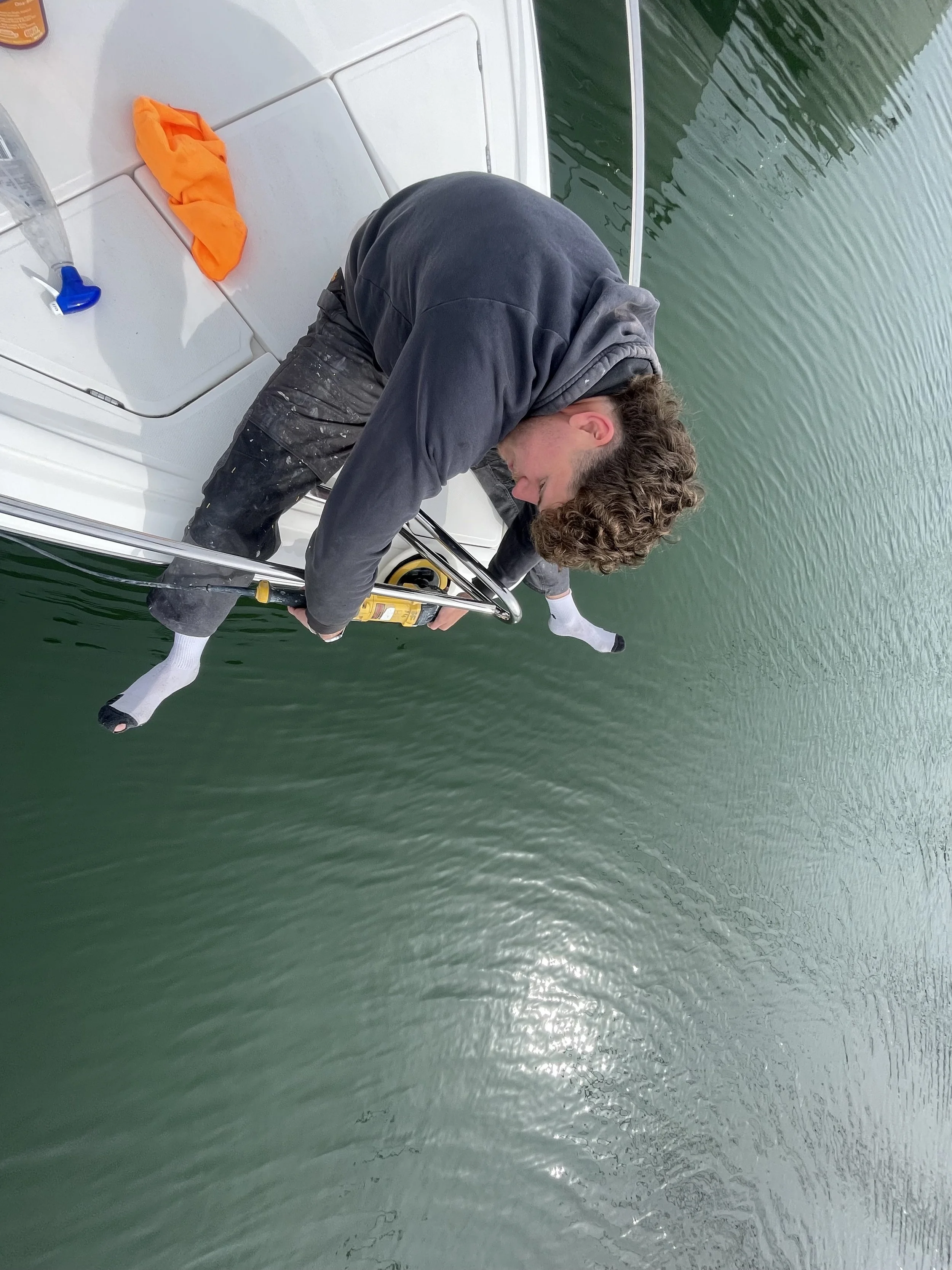 A person with curly hair is leaning over the side of a boat, wearing black clothes and white socks, holding onto a railing and a yellow device, with water below. There are objects on the boat deck, including an orange item and a water bottle.