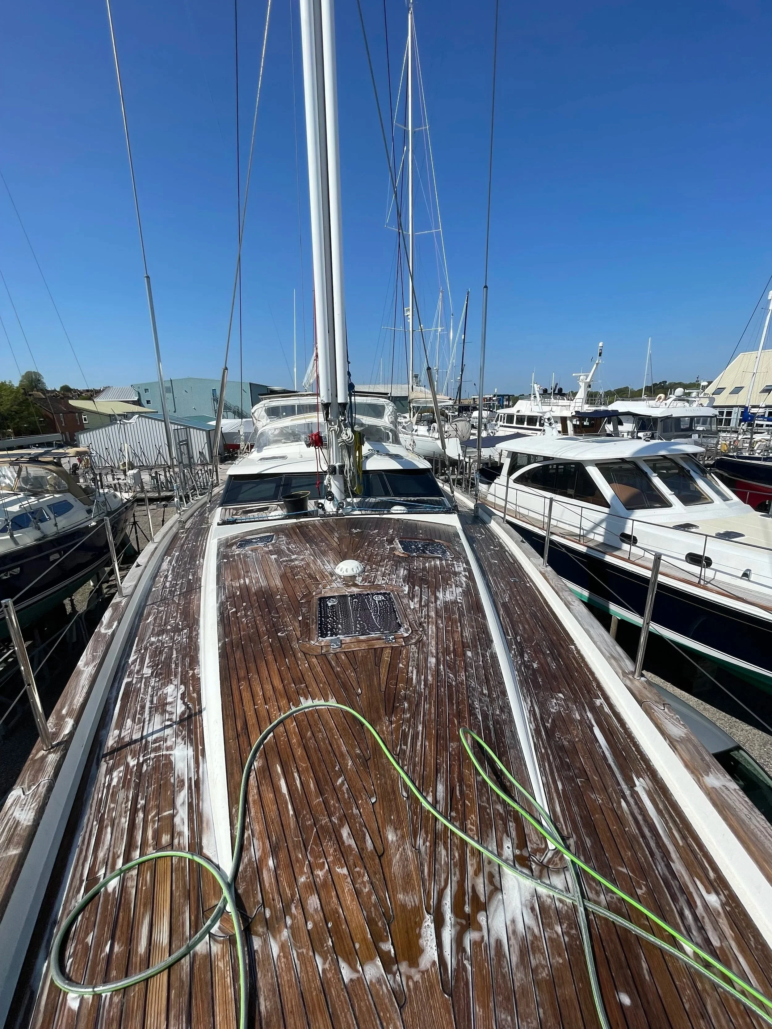 View of a boat docked at a marina with a clear blue sky above. The wooden deck of the boat is wet, with a green hose on it.