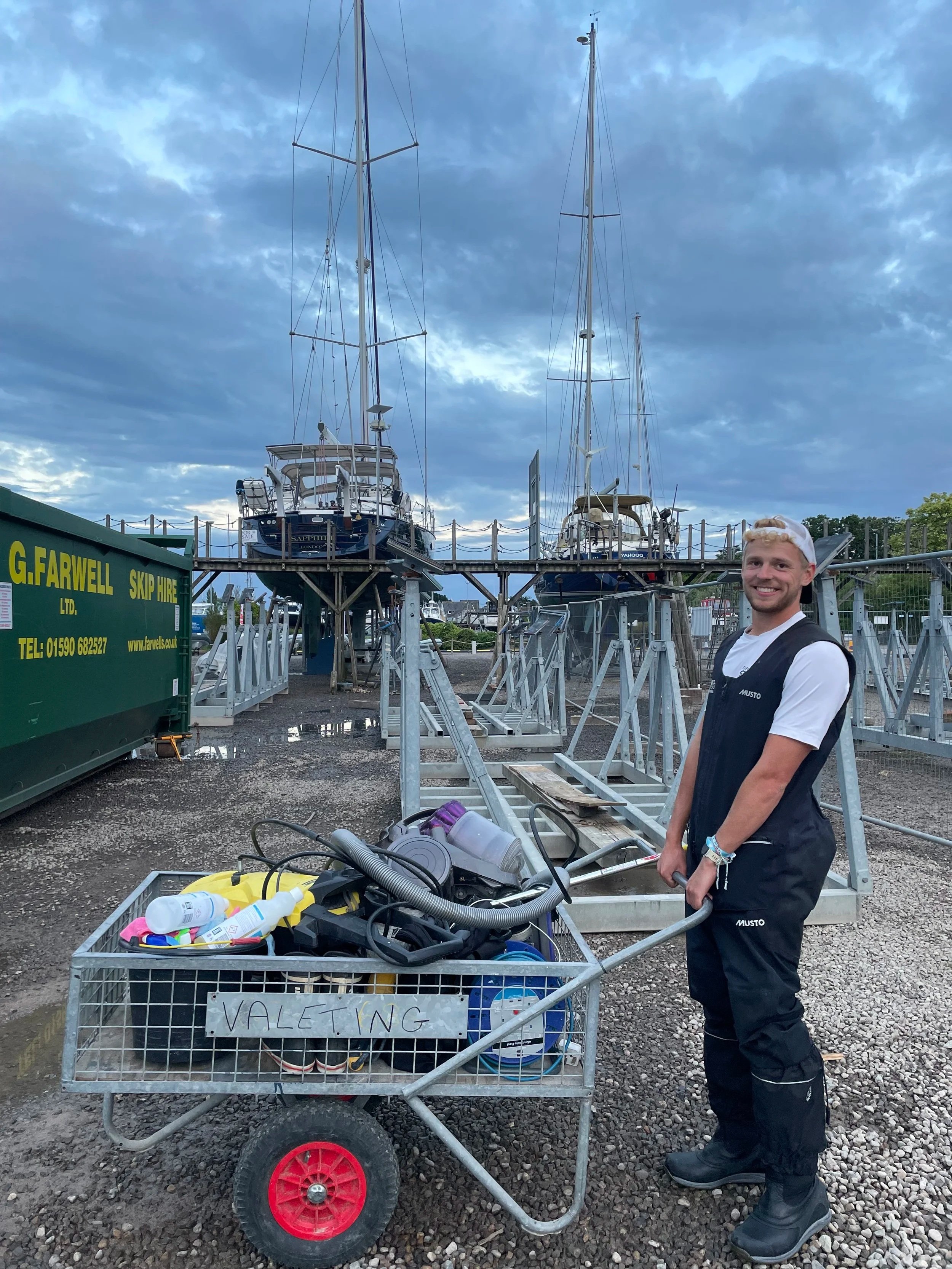 A young man with blonde hair and a white cap smiling at the camera, standing next to a metal cart labeled "Valeting" filled with cleaning supplies, in front of sailboats on a boatyard or marina. The background shows a cloudy sky and sailboats on a lift.