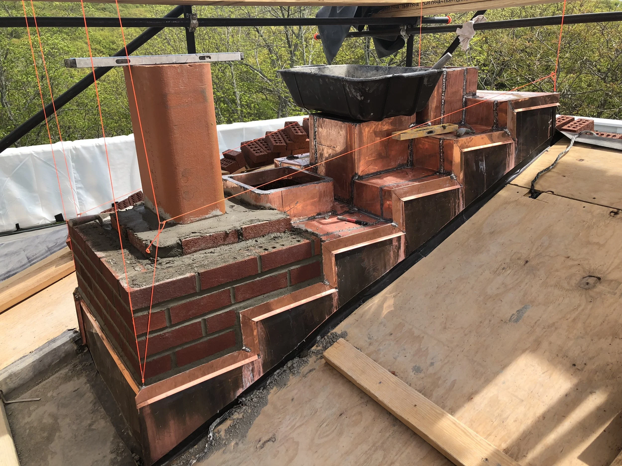 Partially constructed brick chimney on a wooden floor with construction tools and a level placed across the top. Orange safety string lines are stretched across the area for alignment. Green foliage visible in the background.