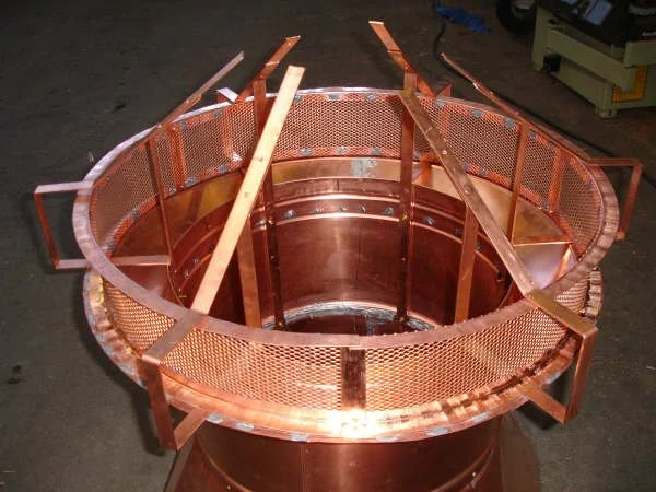 A large, circular copper or bronze metallic structure with a mesh fence around the top edge, supported by vertical beams, placed on a workshop floor.