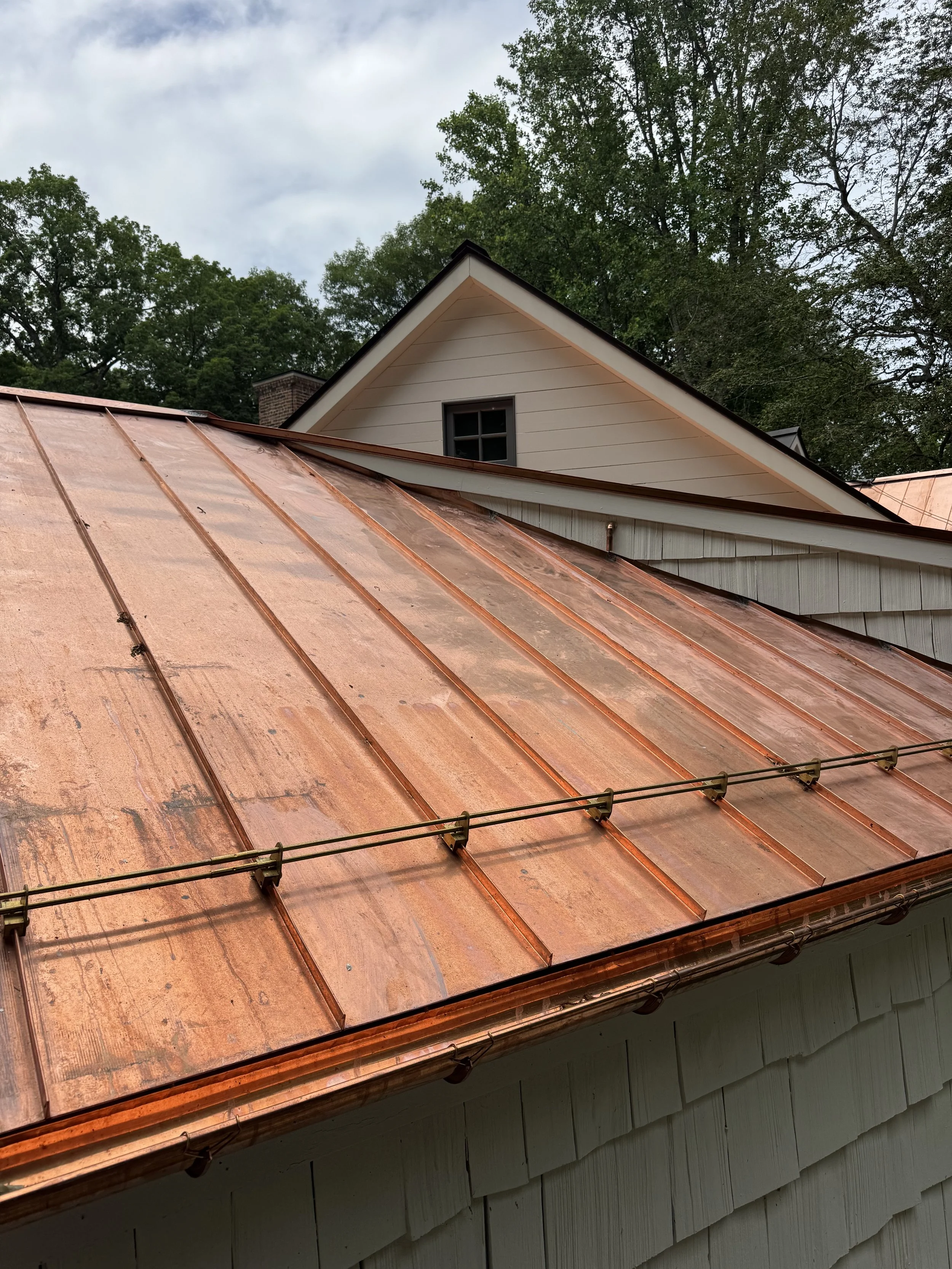 Copper roof shingles on a house with a gable roof and a small window, with trees and a cloudy sky in the background.