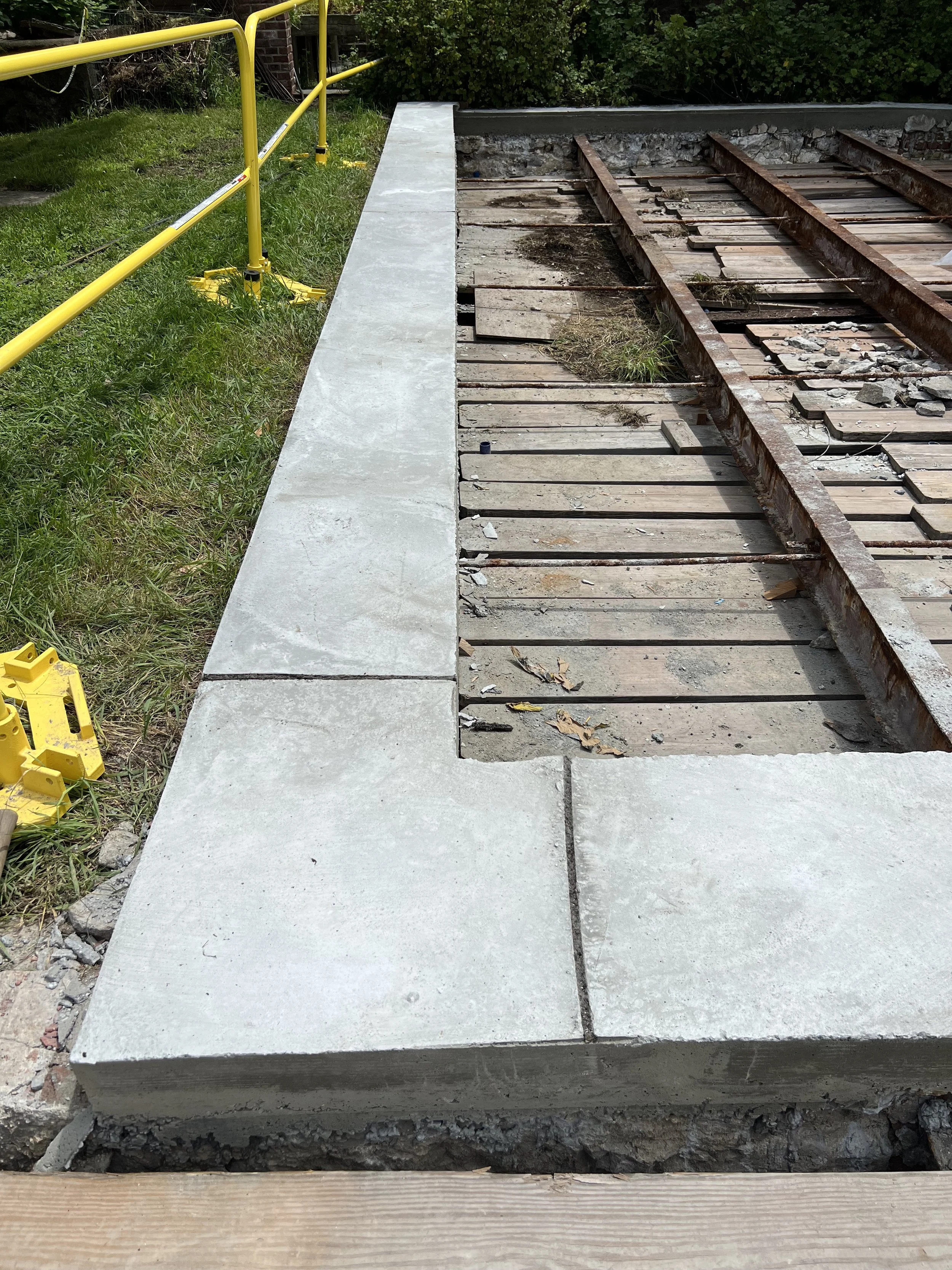 Construction site with a concrete sidewalk and removed railroad tracks, with yellow safety barriers and green grass on the side.