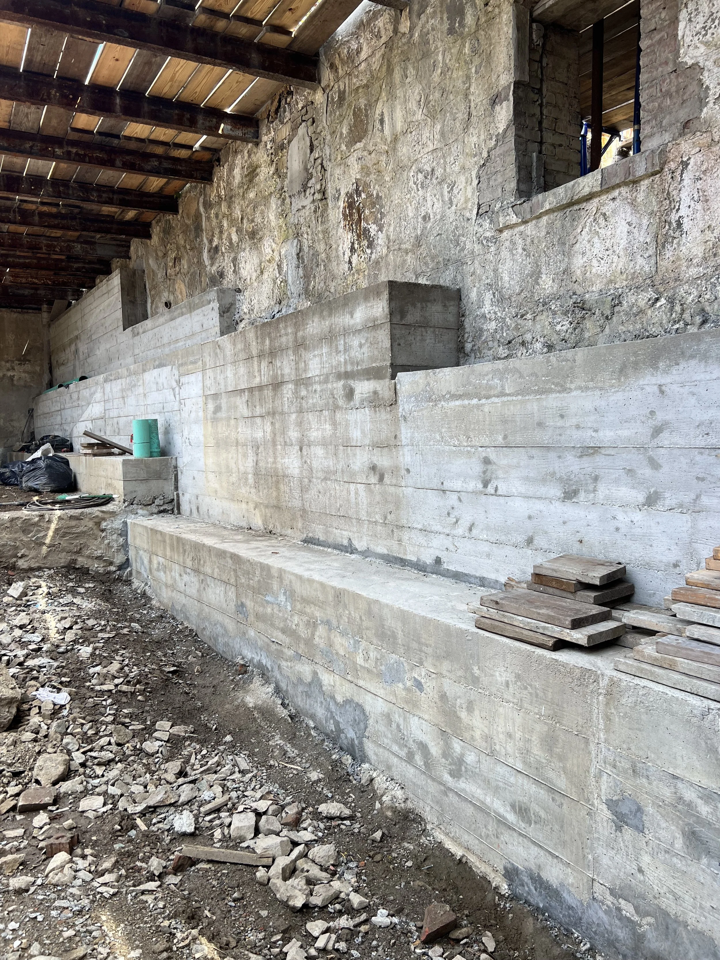 Construction site with concrete foundation and rocky wall, scattered rocks and tools on the ground, and wooden roofing above.