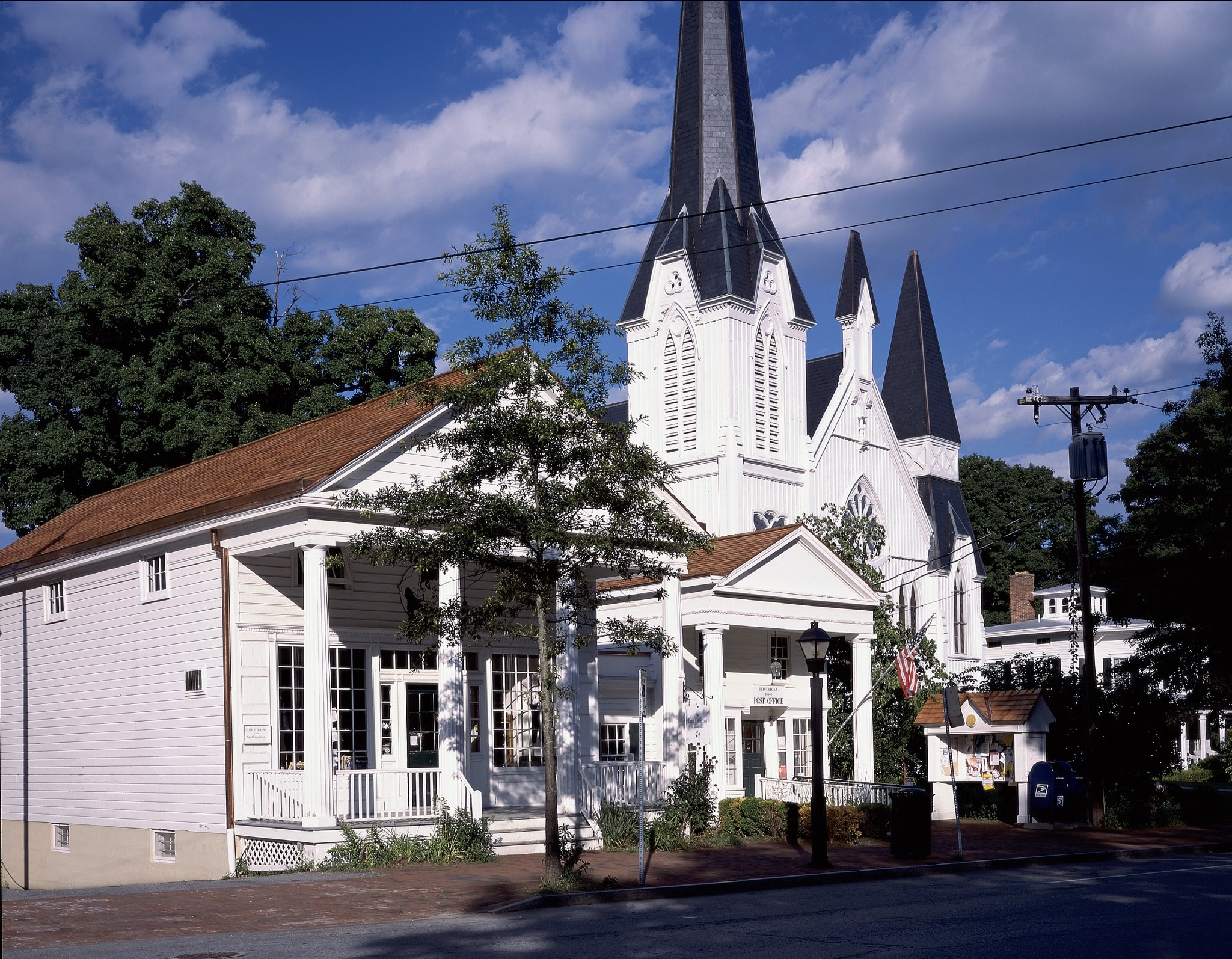 A white church with black steeples and a red roof, with trees and power lines in front of it on a partly cloudy day.