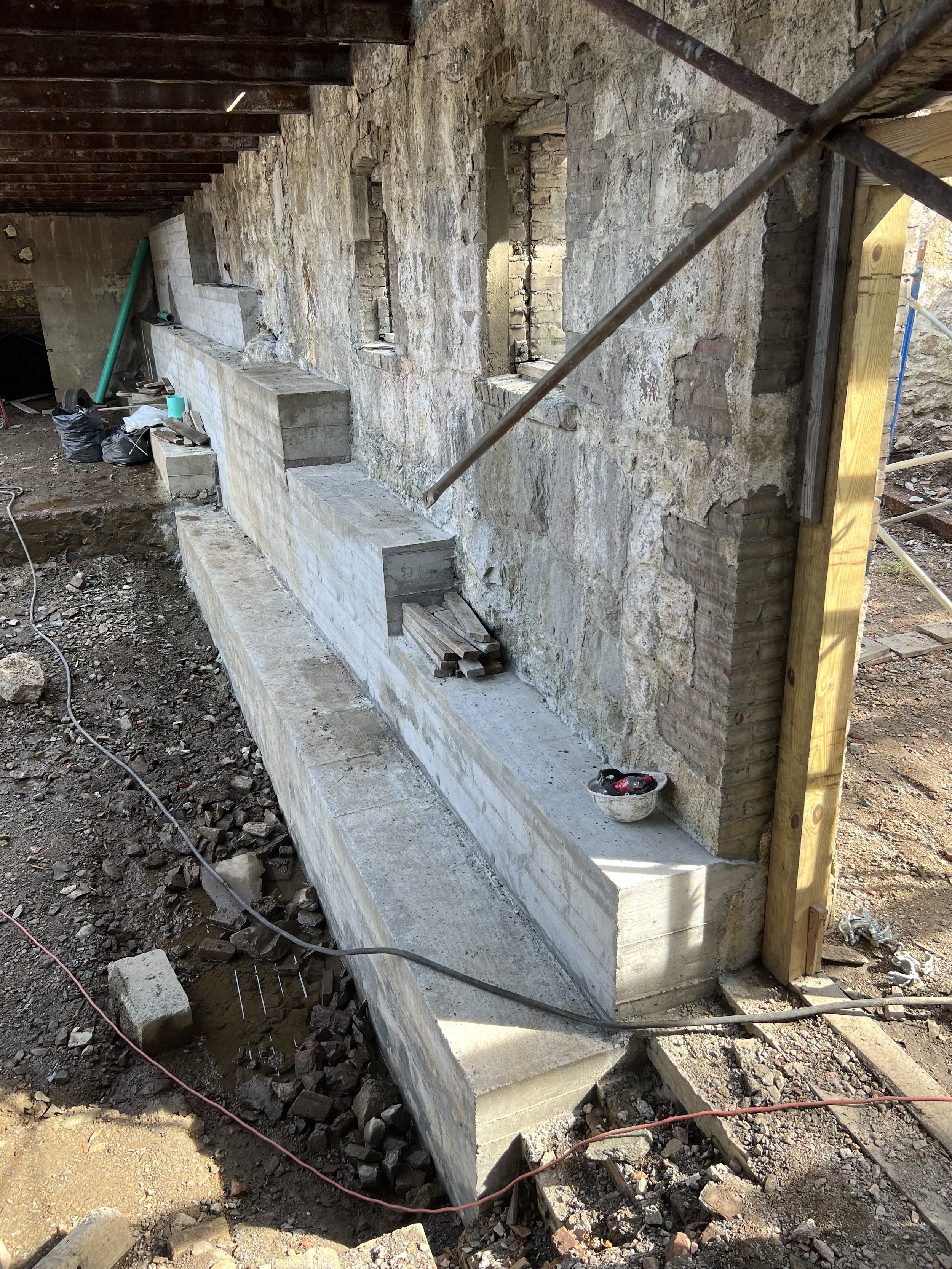 Construction site showing a stone wall under renovation with a concrete ledge and exposed brickwork. Building tools and materials are scattered around, with a wooden and metal scaffolding structure on the side.