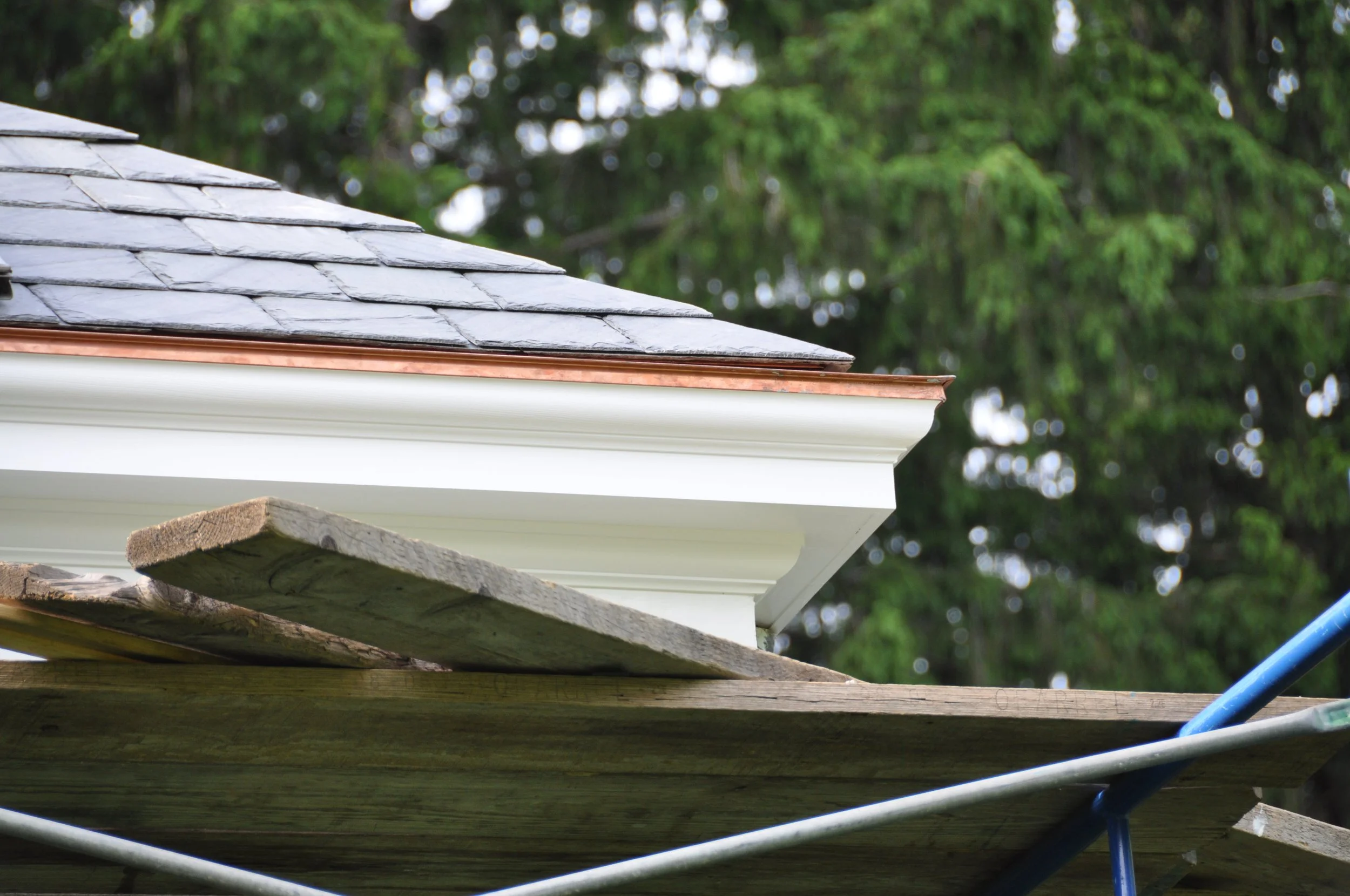 Close-up of a house roof with slate shingles, copper flashing, and white siding, with green trees in the background.